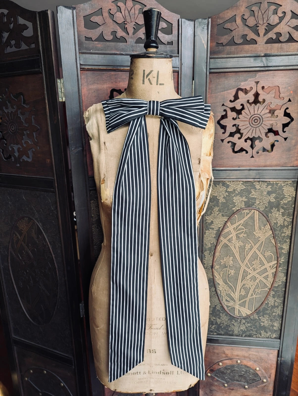 Broochella large, cotton, handmade black and white striped  bow. It is pinned to a mannequin. The background is a decorative brown wooden screen.