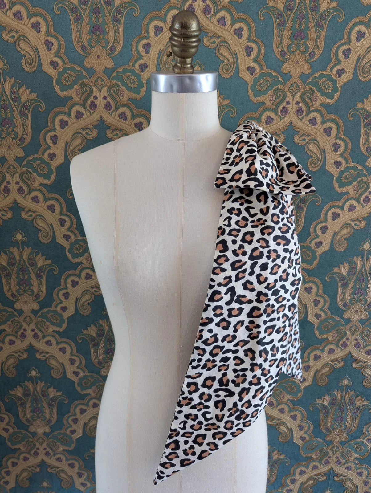 Broochella large, cotton, handmade leopard pint bow. It is pinned to a mannequin. The background is green and gold damask wallpaper.