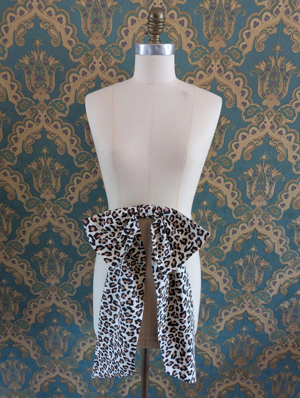Broochella large, cotton, handmade leopard pint bow. It is pinned to a mannequin. The background is green and gold damask wallpaper.