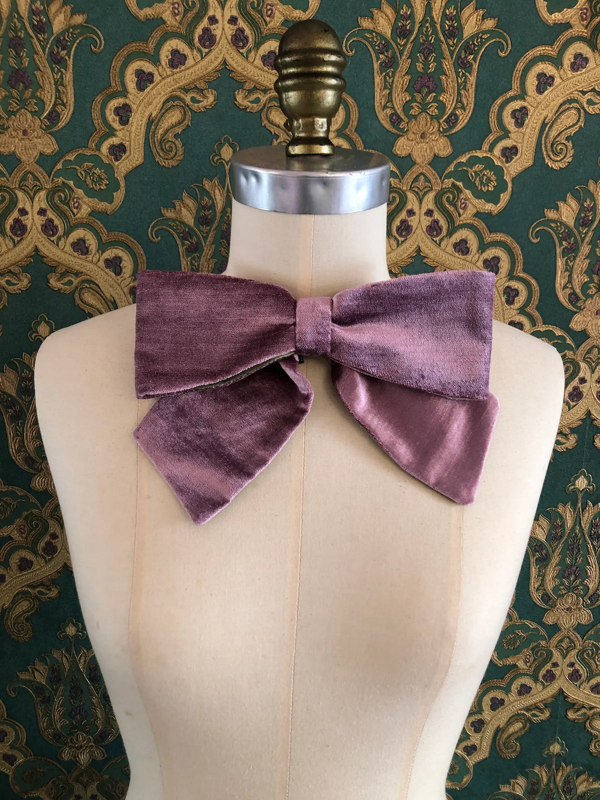 Broochella handmade Large Velvet Bow Brooch in soft violet velvet with contrasting green lining, styled asymmetrically on a cream mannequin against ornate green and gold wallpaper.