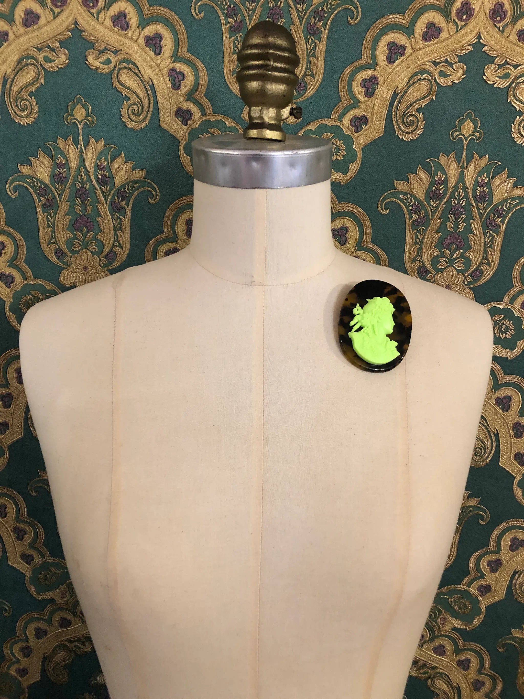 Broochella La Bella Figura Cameo brooch. A lime cameo on a tortoiseshell base,  displayed on a cream dress form against a dark teal and gold patterned fabric backdrop.