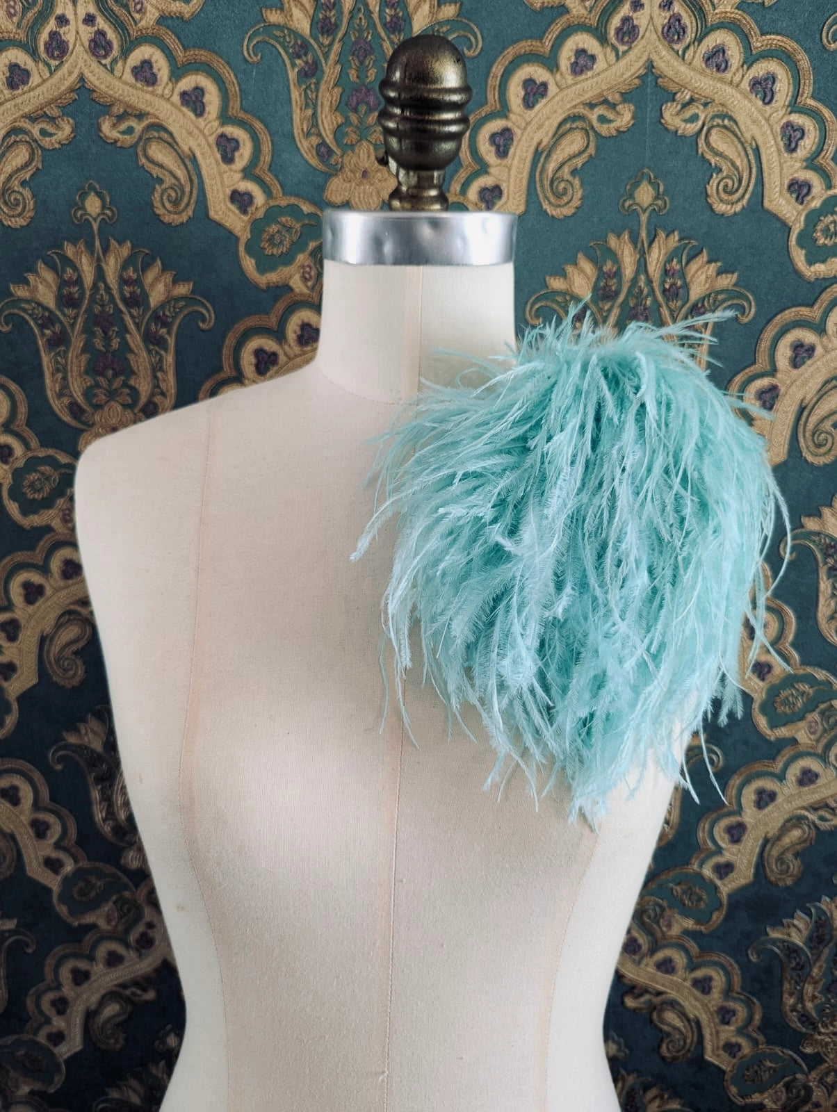 Broochella le pouffe handmade designer feather brooch in aqua.  It is pinned to a mannequin. The background is green and gold damask wallpaper