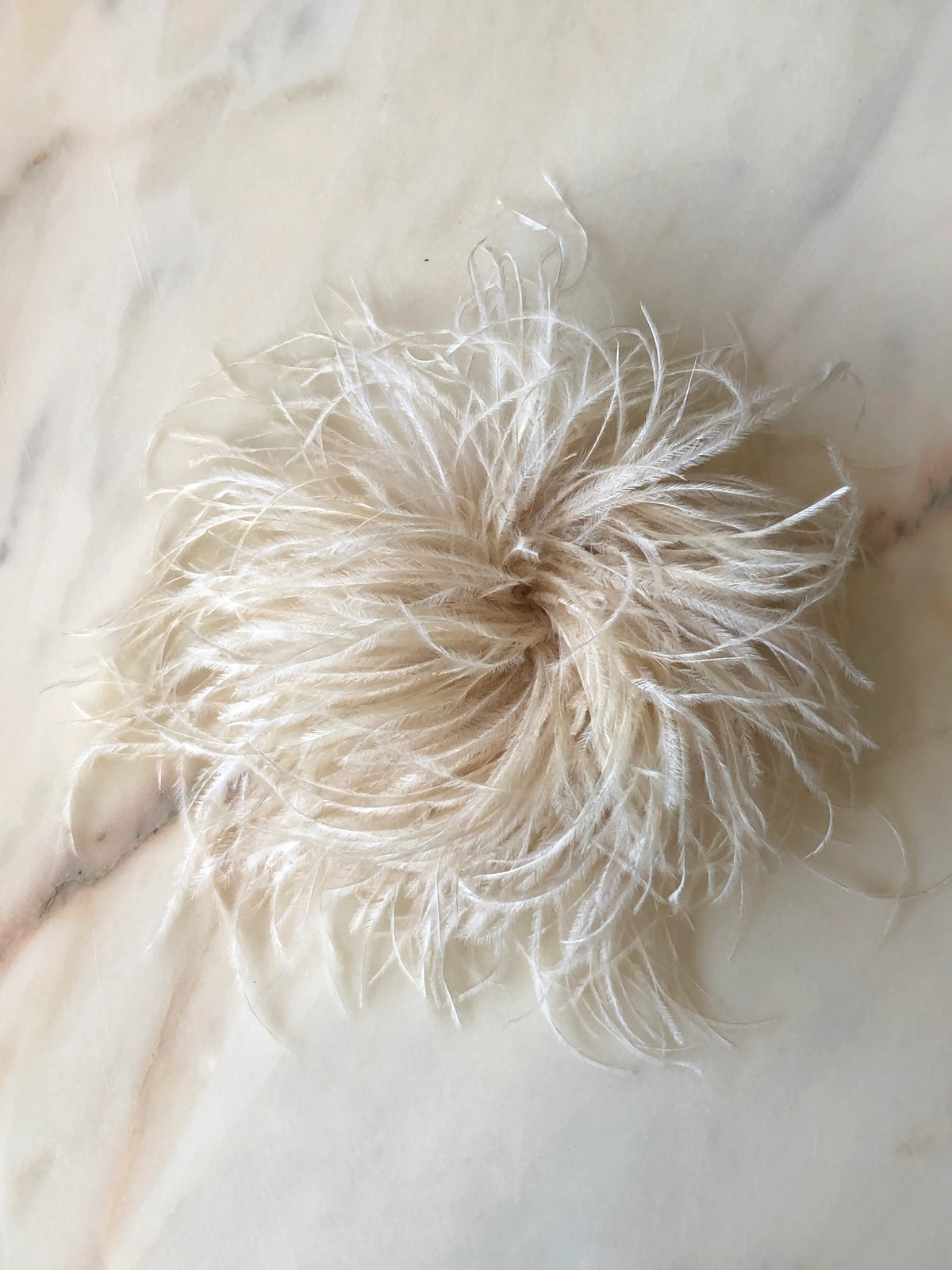 Broochella Le Pouffe Feather Brooch in soft beige, featuring a voluminous arrangement of wispy ostrich feathers, photographed on a pale white marble background with subtle grey and blush veining.