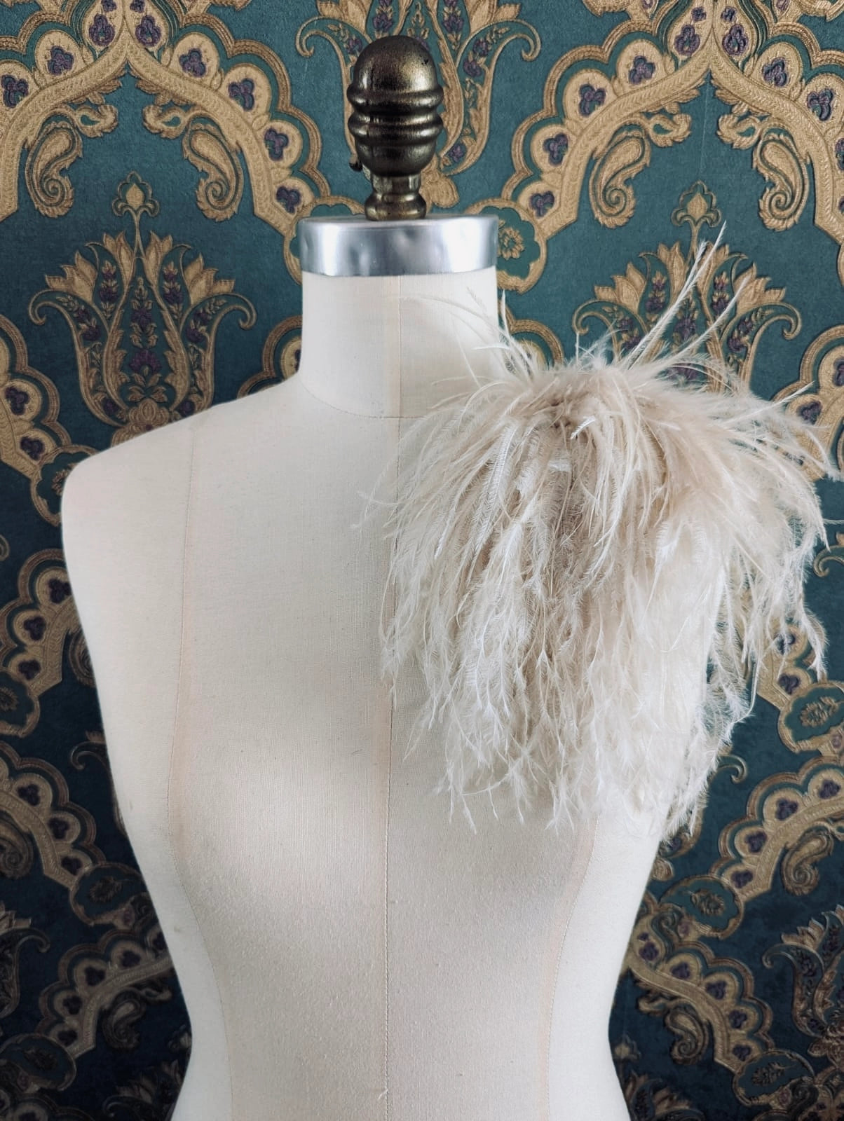 Broochella le pouffe handmade designer feather brooch in beige.  It is pinned to a mannequin. The background is green and gold damask wallpaper