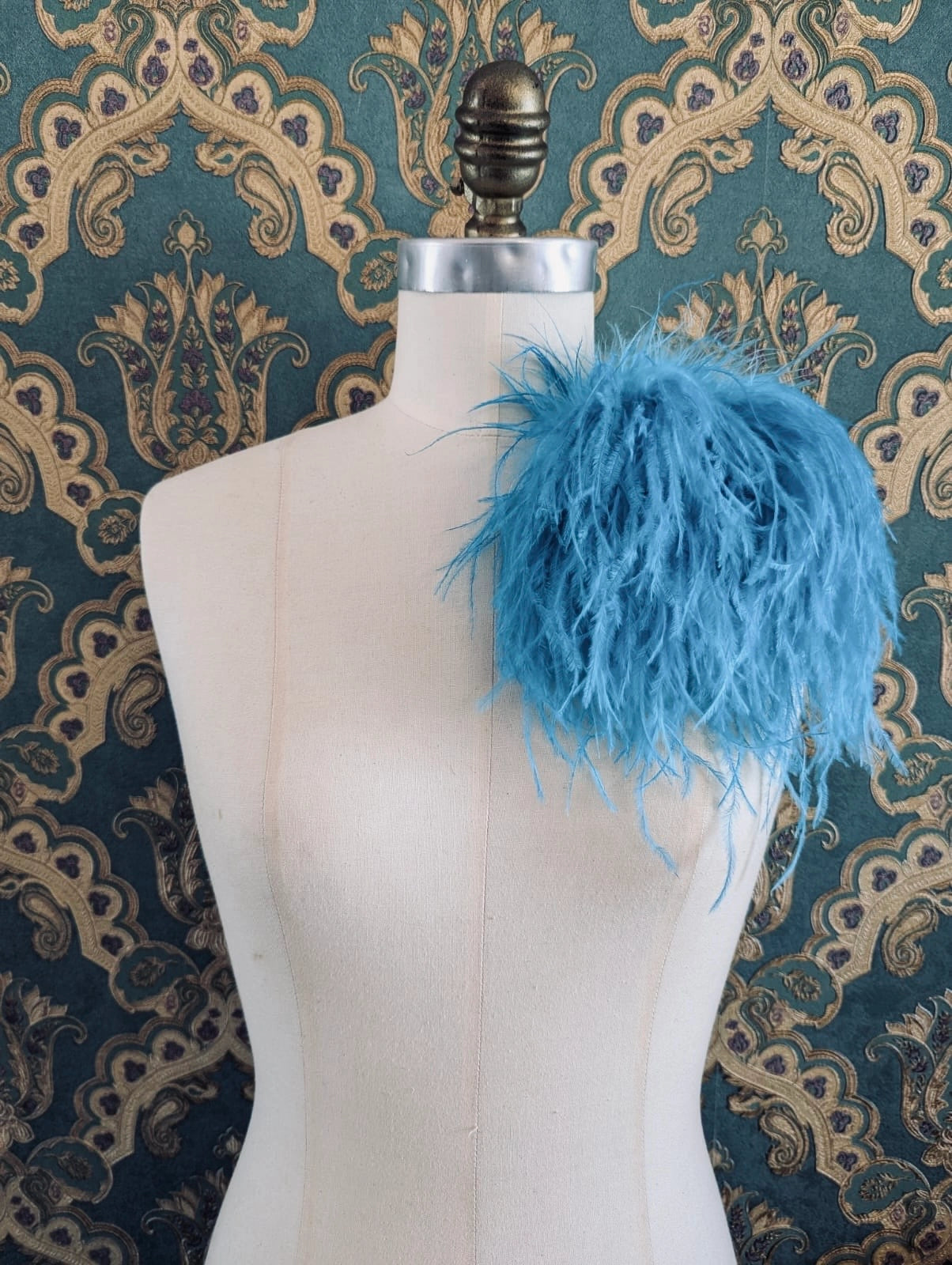 Broochella le pouffe handmade designer feather brooch in blue.  It is pinned to a mannequin. The background is green and gold damask wallpaper