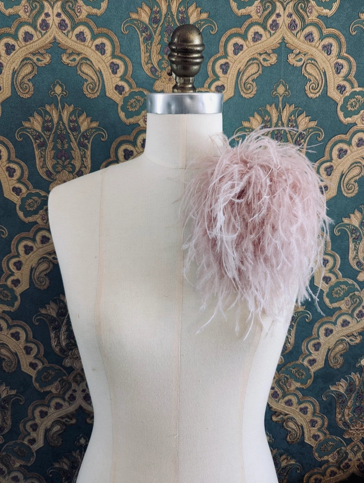 Broochella le pouffe handmade designer feather brooch in blush pink.  It is pinned to a mannequin. The background is green and gold damask wallpaper