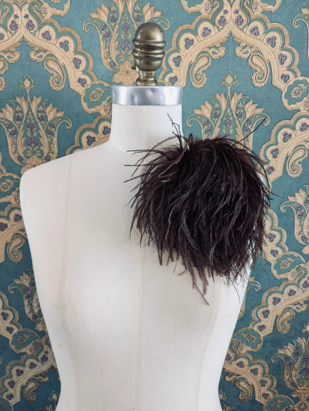 Broochella le pouffe handmade designer feather brooch in brown.  It is pinned to a mannequin. The background is green and gold damask wallpaper