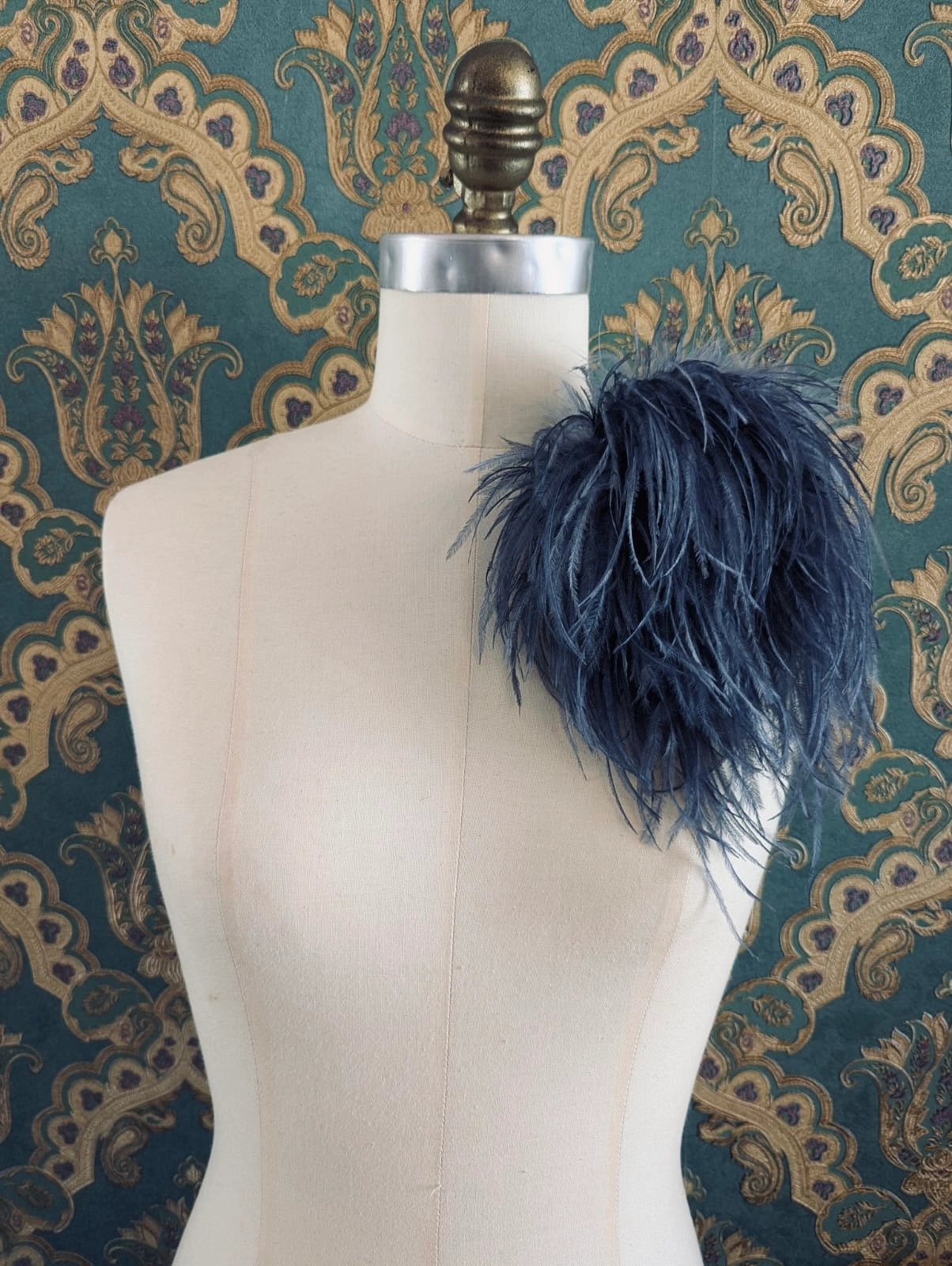 Broochella le pouffe handmade designer feather brooch in grey.  It is pinned to a mannequin. The background is green and gold damask wallpaper.