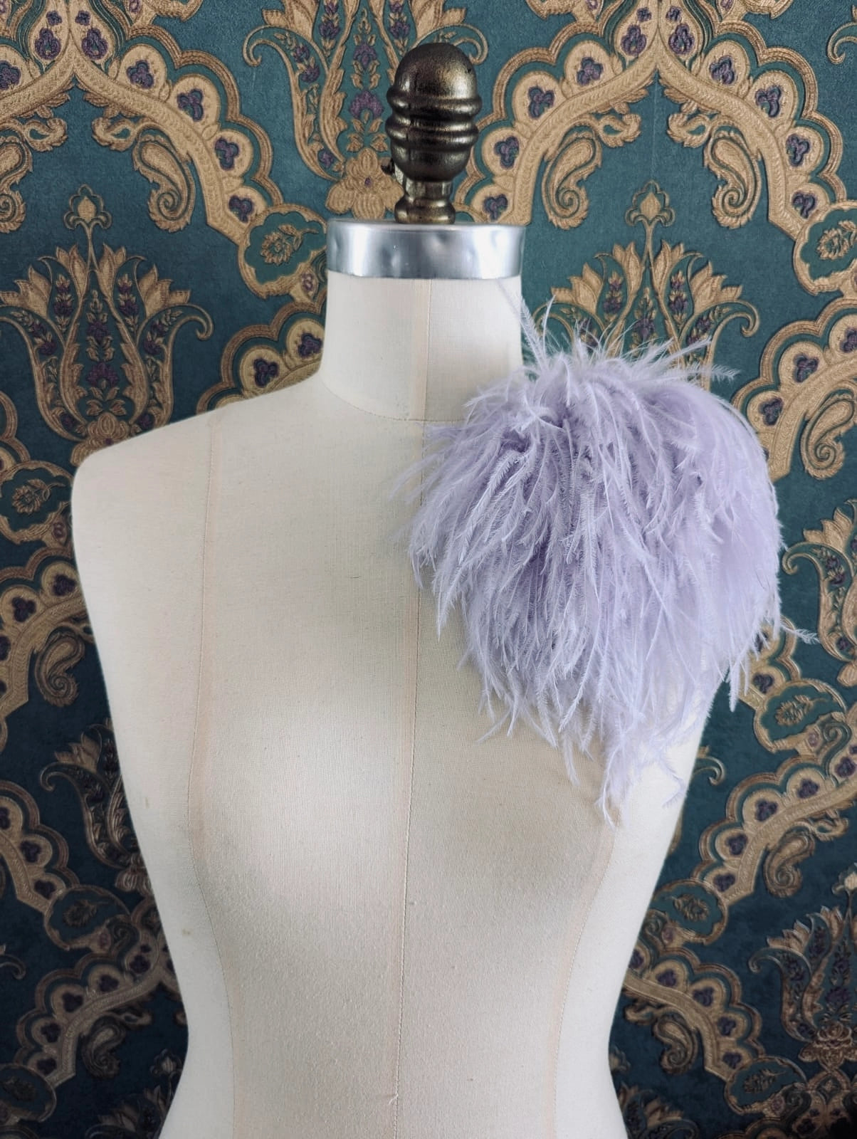 Broochella le pouffe handmade designer feather brooch in lilac.  It is pinned to a mannequin. The background is green and gold damask wallpaper