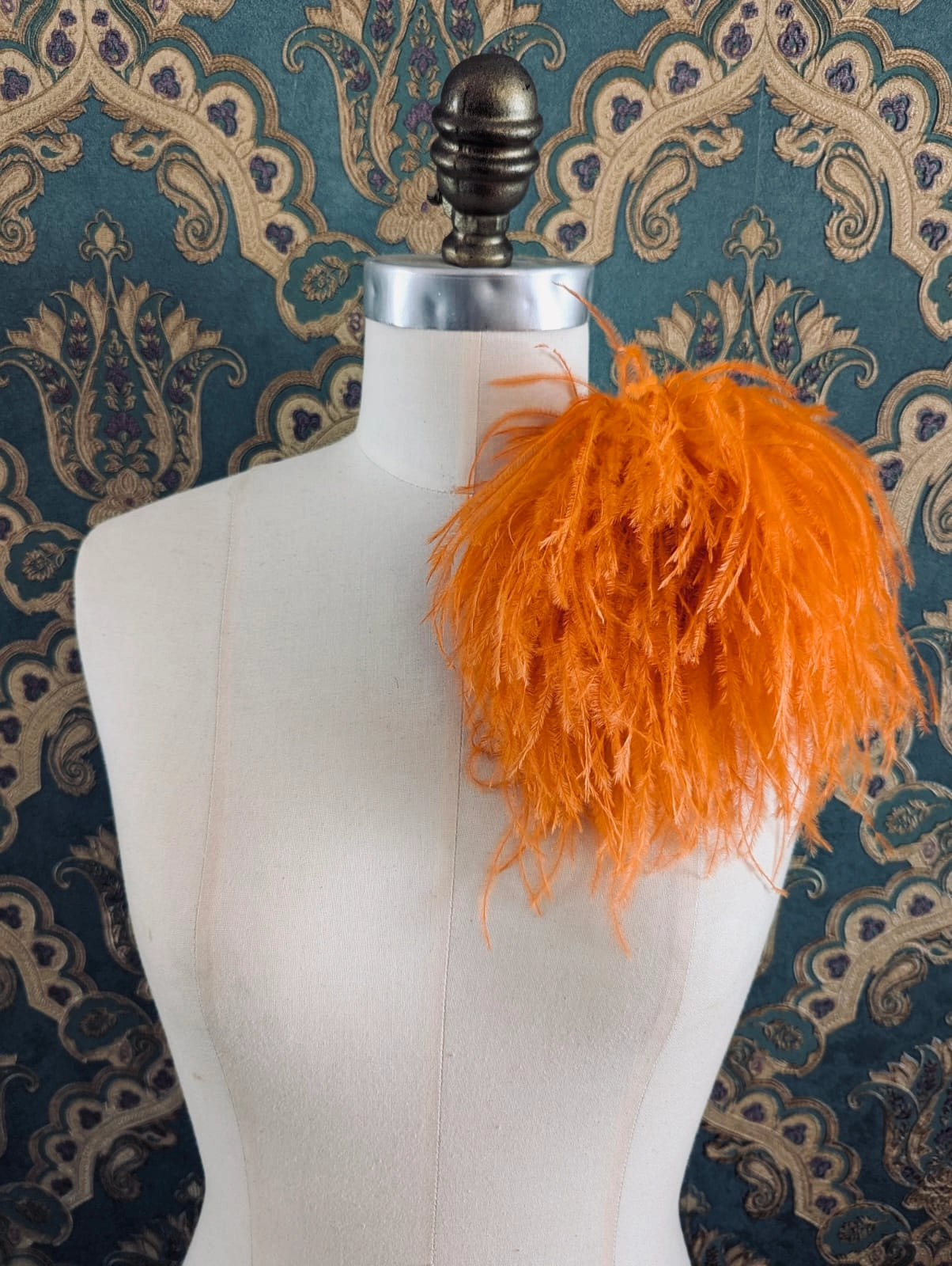 Broochella le pouffe handmade designer feather brooch in orange.  It is pinned to a mannequin. The background is green and gold damask wallpaper