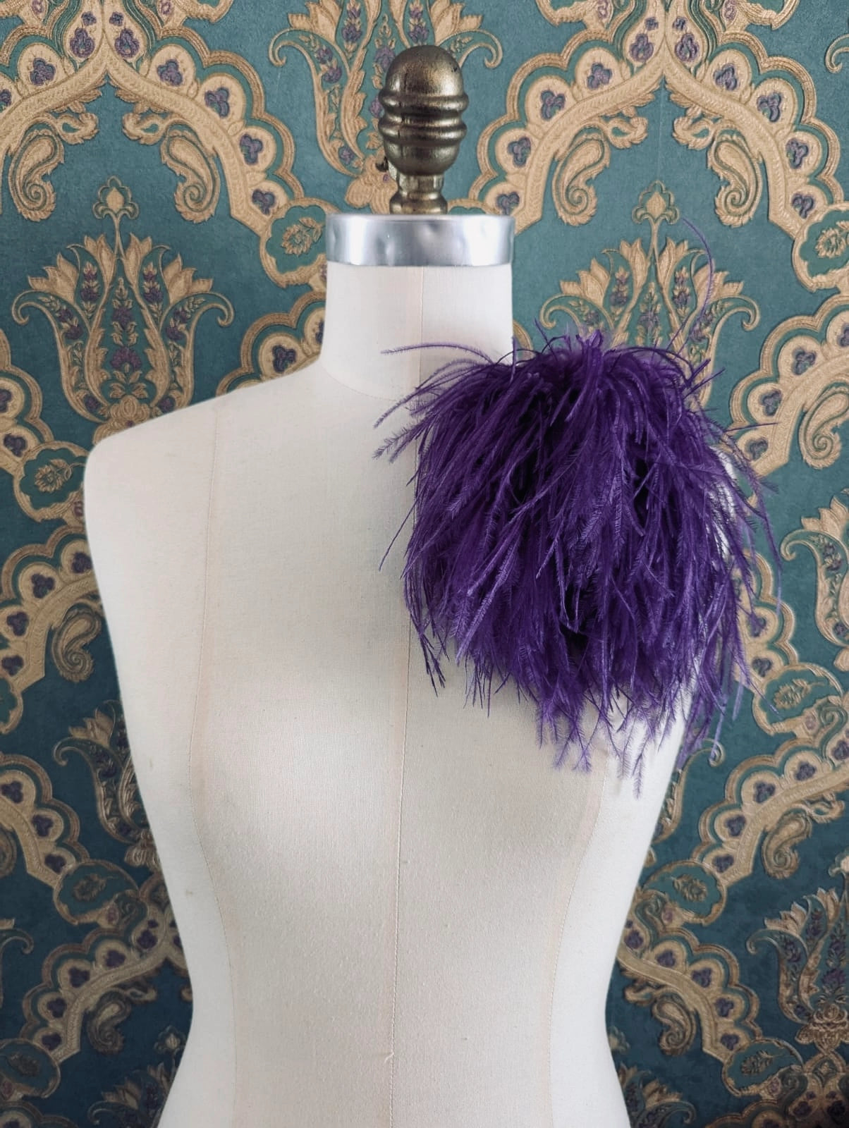 Broochella le pouffe handmade designer feather brooch in purple.  It is pinned to a mannequin. The background is green and gold damask wallpaper