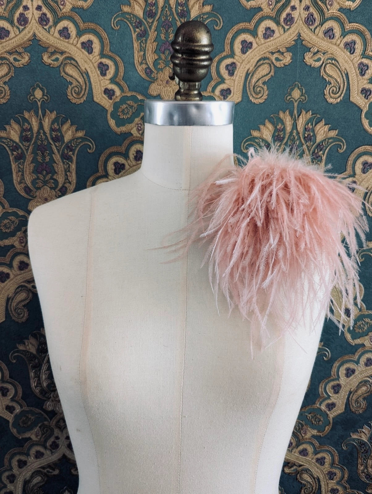 Broochella le pouffe handmade designer feather brooch in rose gold.  It is pinned to a mannequin. The background is green and gold damask wallpaper