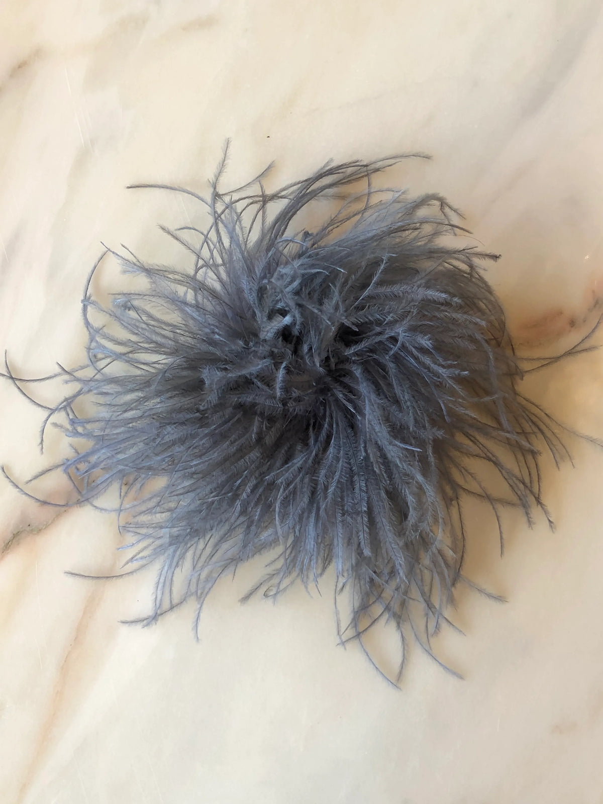 Broochella Le Pouffe Feather Brooch in silver grey, featuring a voluminous arrangement of soft, wispy feathers, photographed on a pale white marble background with subtle grey and blush veining.