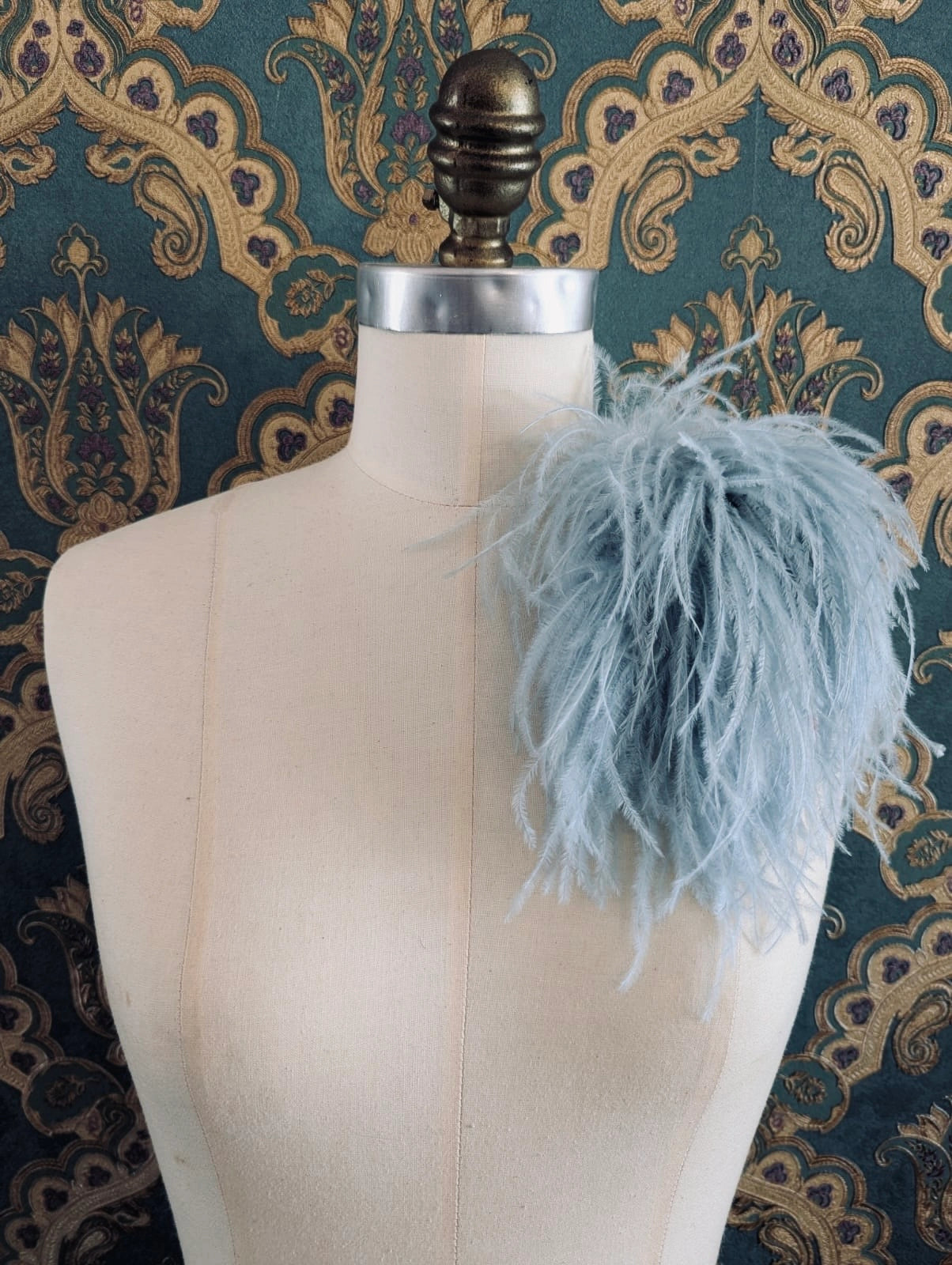 Broochella le pouffe handmade designer feather brooch in sky blue.  It is pinned to a mannequin. The background is green and gold damask wallpaper