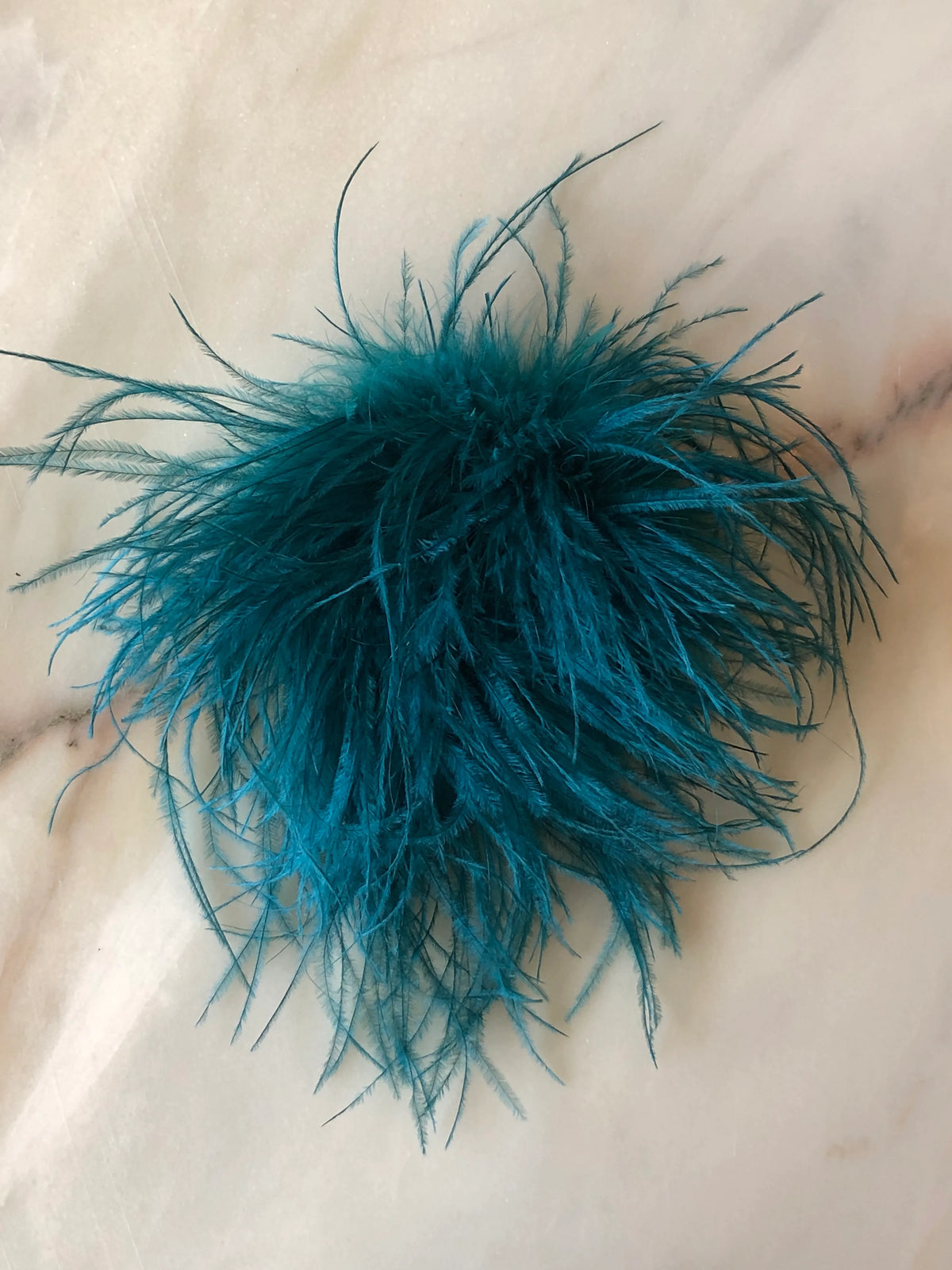 Broochella Le Pouffe Feather Brooch in rich teal, featuring a voluminous arrangement of soft, wispy feathers, photographed on a pale white marble background with subtle grey and blush veining.