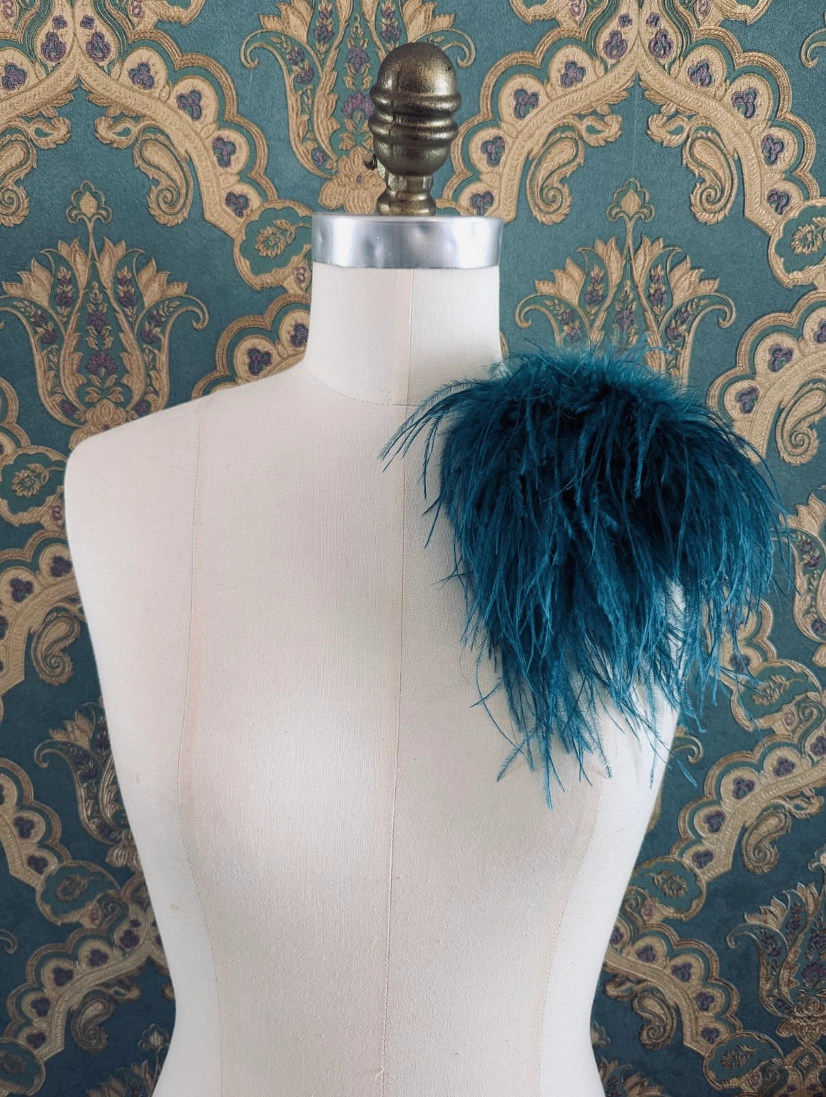 Broochella le pouffe handmade designer feather brooch in teal.  It is pinned to a mannequin. The background is green and gold damask wallpaper