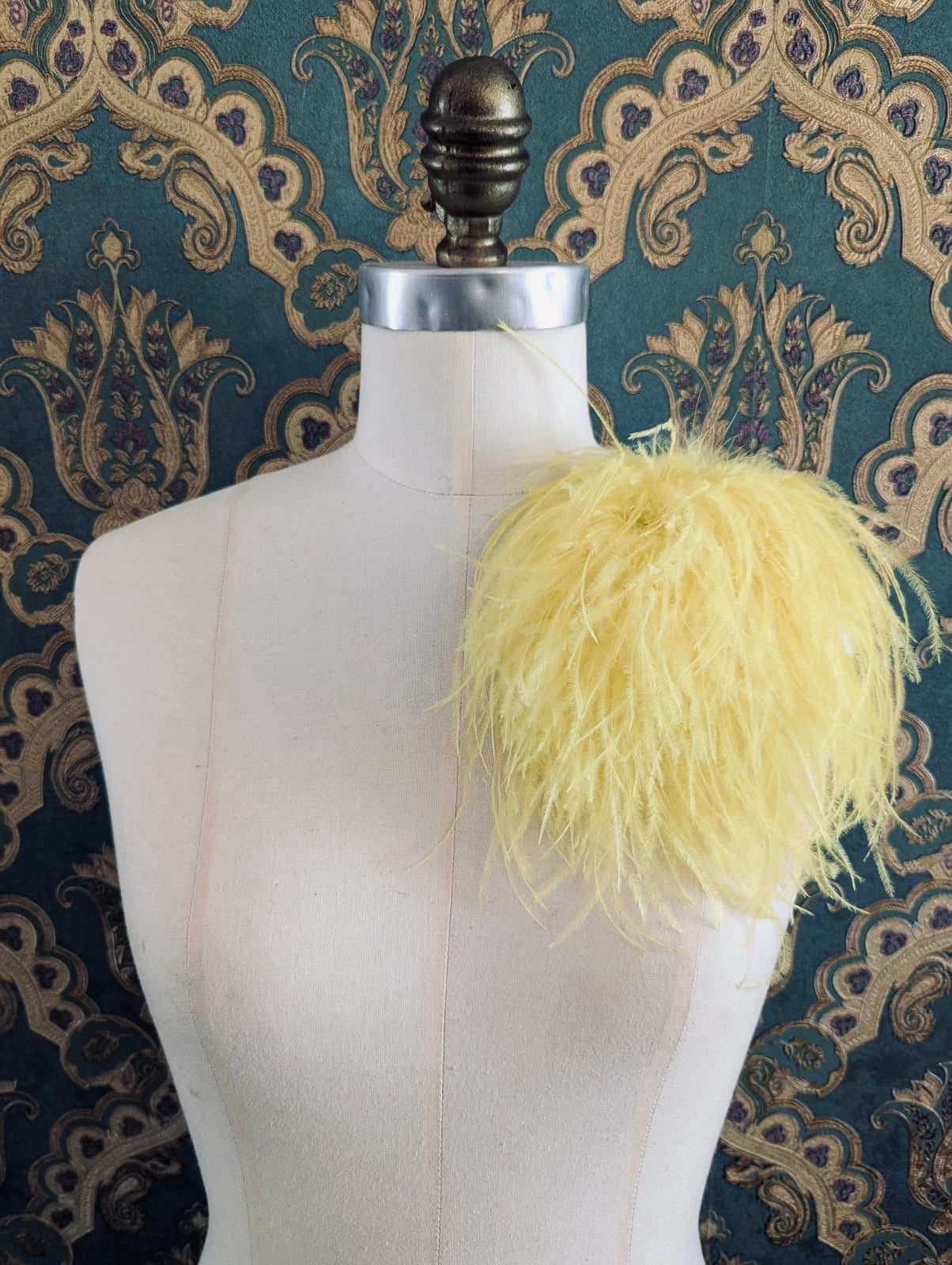 Broochella le pouffe handmade designer feather brooch in yellow.  It is pinned to a mannequin. The background is green and gold damask wallpaper