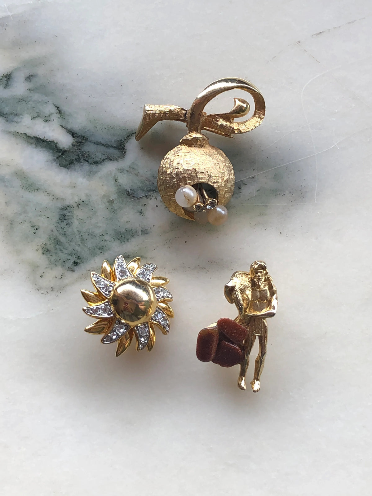 Broochella Les Petites Curiosités Vintage Brooch Trio featuring a textured gold-tone bell with faux pearls, a gold and clear crystal flower, and a miniature gold figure with brown accents, photographed on a white marble background with soft grey-green veining.