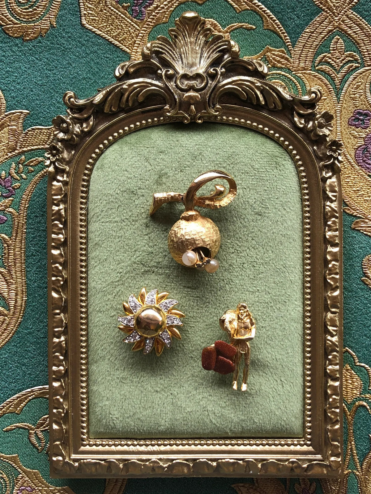 Broochella Les Petites Curiosités Vintage Brooch Trio featuring three whimsical gold-tone brooches, including a pearl-detailed bell, crystal flower and miniature figure with brown accents, displayed on sage green velvet in an ornate antique gold frame against green and gold patterned fabric.