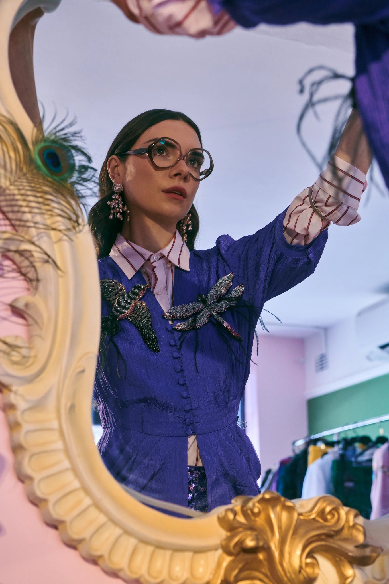 A woman wearing a purple textured jacket with Broochella Libellula dragonfly brooch embellishments, pink and white striped gloves, and round glasses, looking into an ornate gold mirror in a boutique.