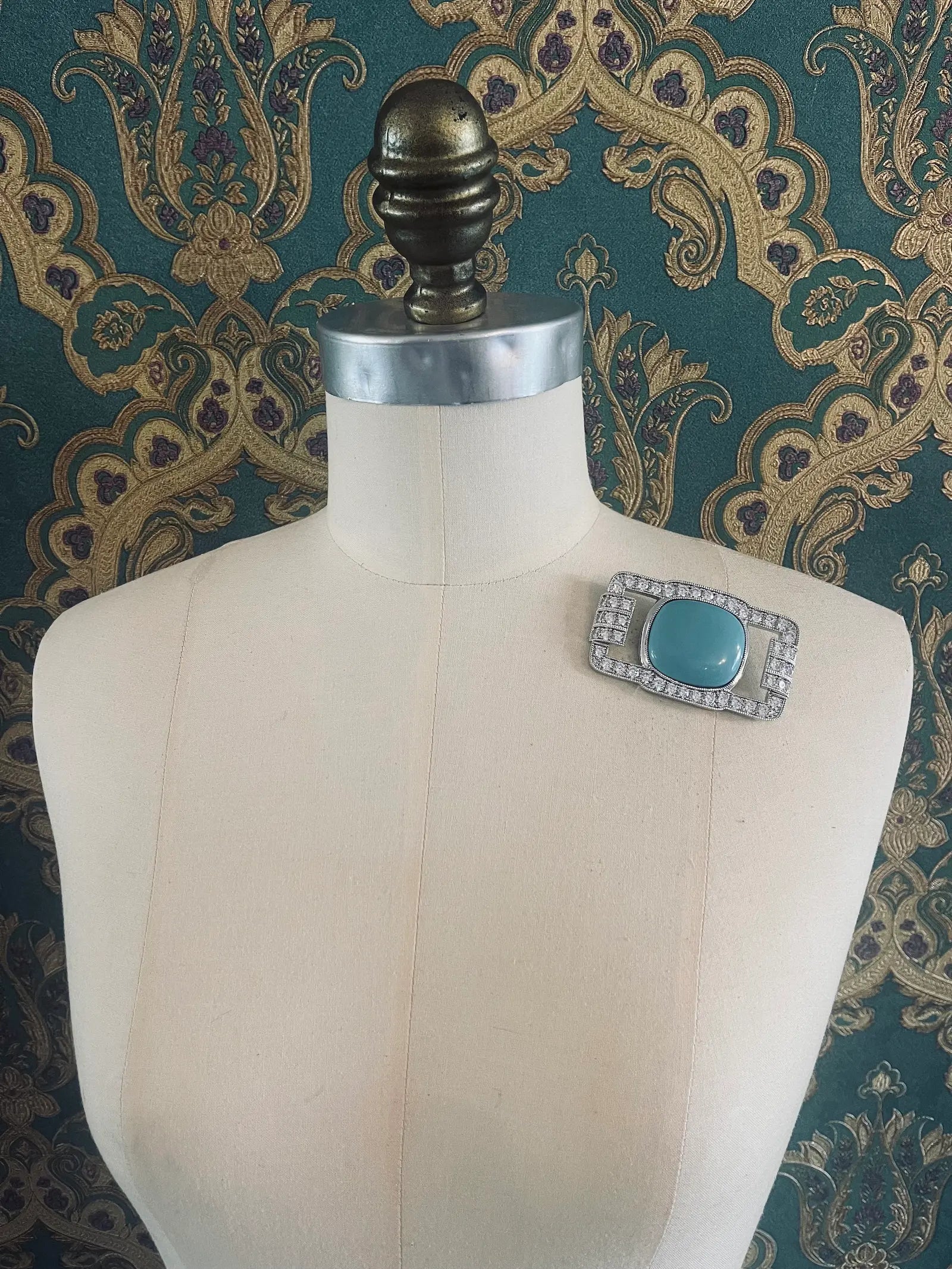 Broochella Libertenè designer silver metal, crystal and turquoise brooch. Pictured on a mannequin. The background is green and gold damask wallpaper