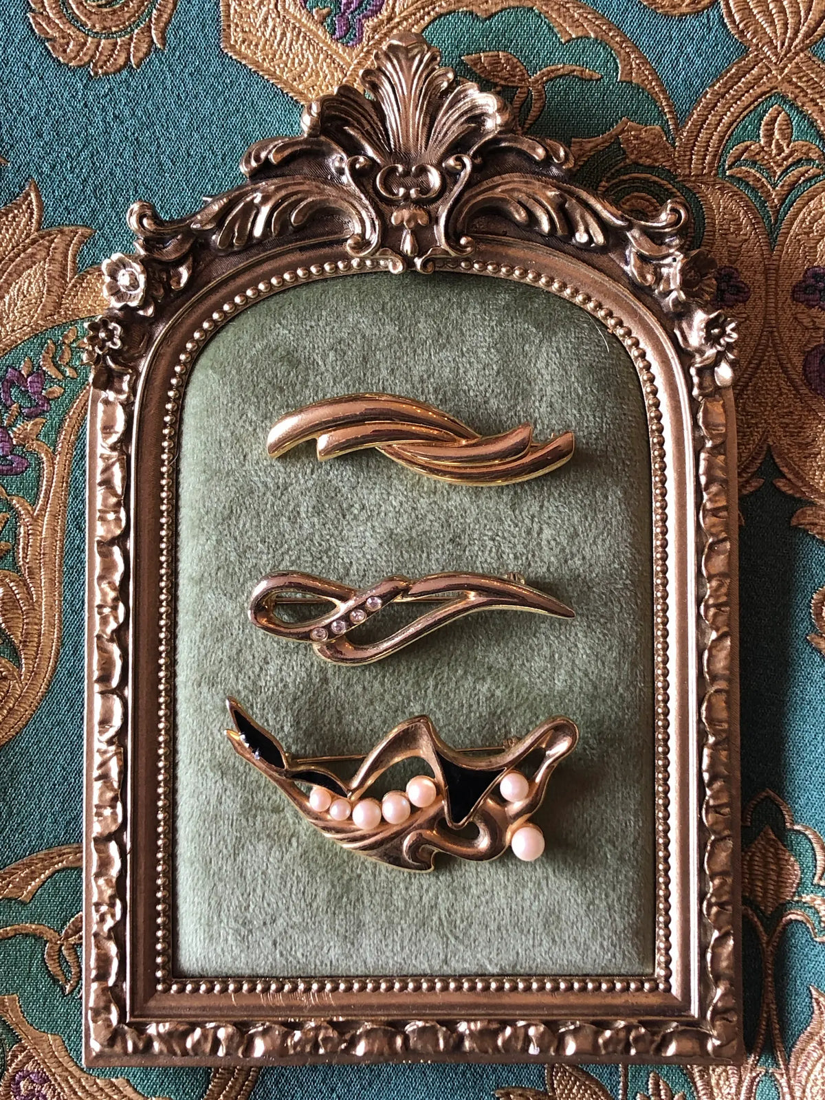 Broochella Linea Lucente vintage brooch set featuring three sculptural gold-tone brooches displayed in an ornate gold frame on sage green velvet, against a teal and gold embroidered fabric background.
