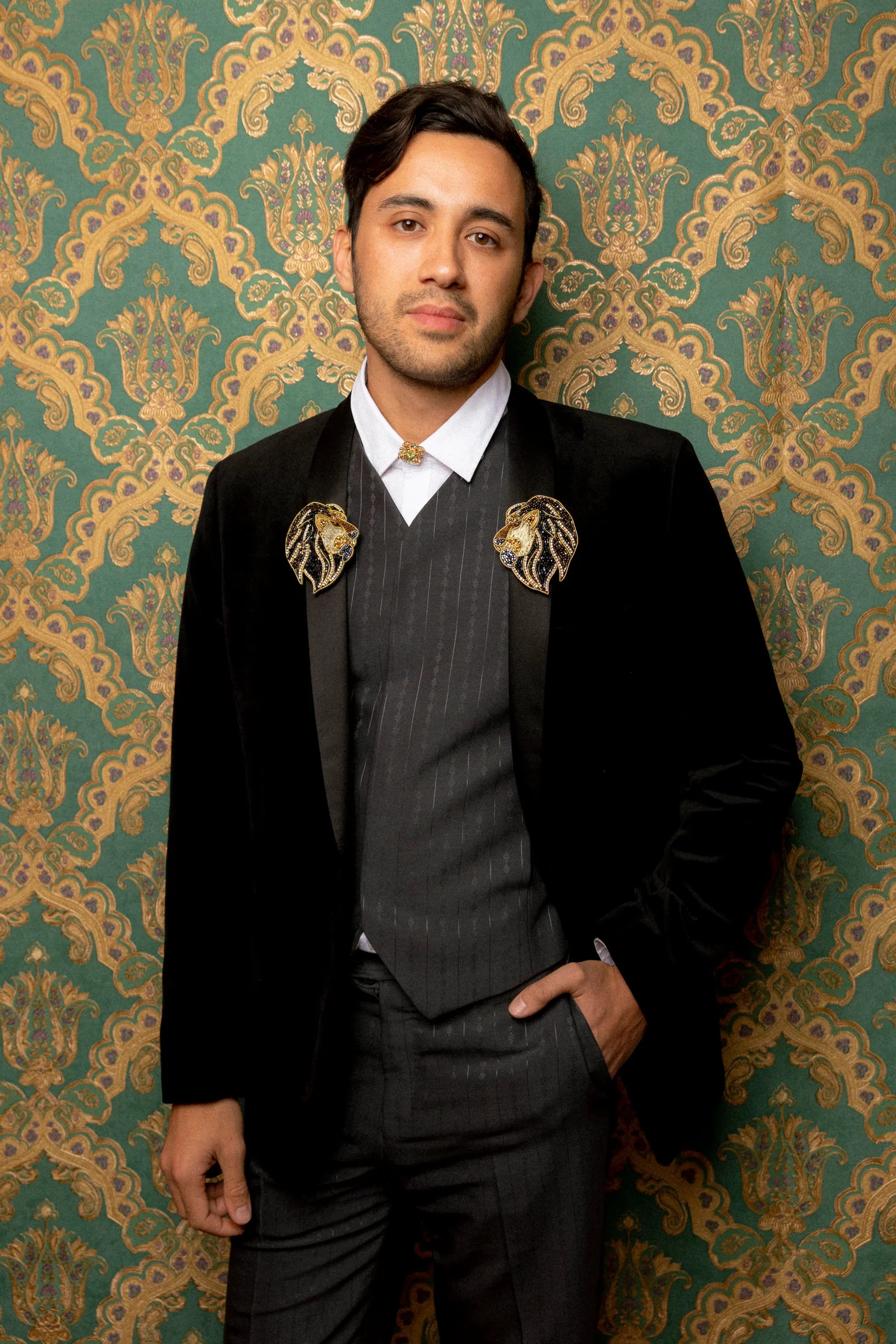 Broochella lion brooches on a male model in a grey pinstripe suit. The background is green and gold damask wallpaper