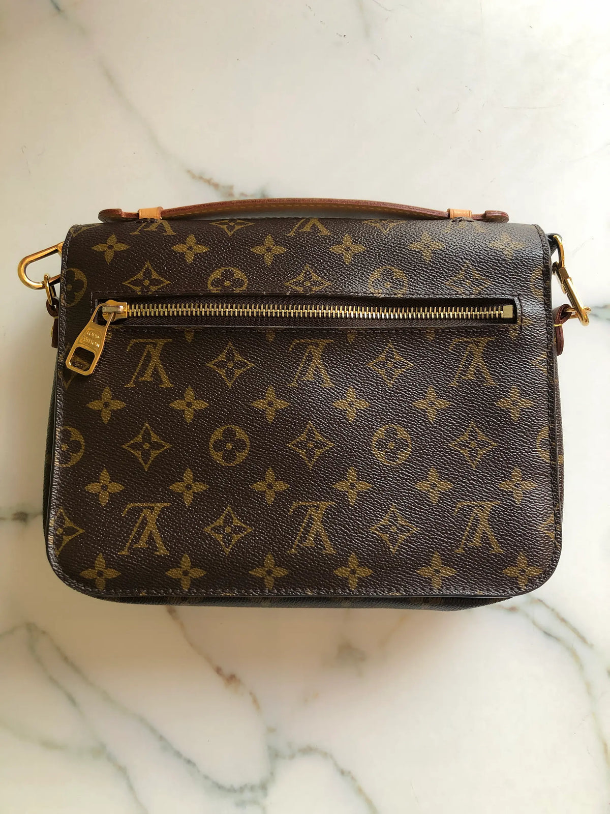 The back view of A Broochella Louis Vuitton Pochette Metis monogram shoulder bag in brown coated canvas with gold-tone hardware, resting on a white marble surface. The bag features the classic LV monogram pattern, a top flap with a gold-tone padlock closure, and an adjustable black leather shoulder strap. The background is white marble