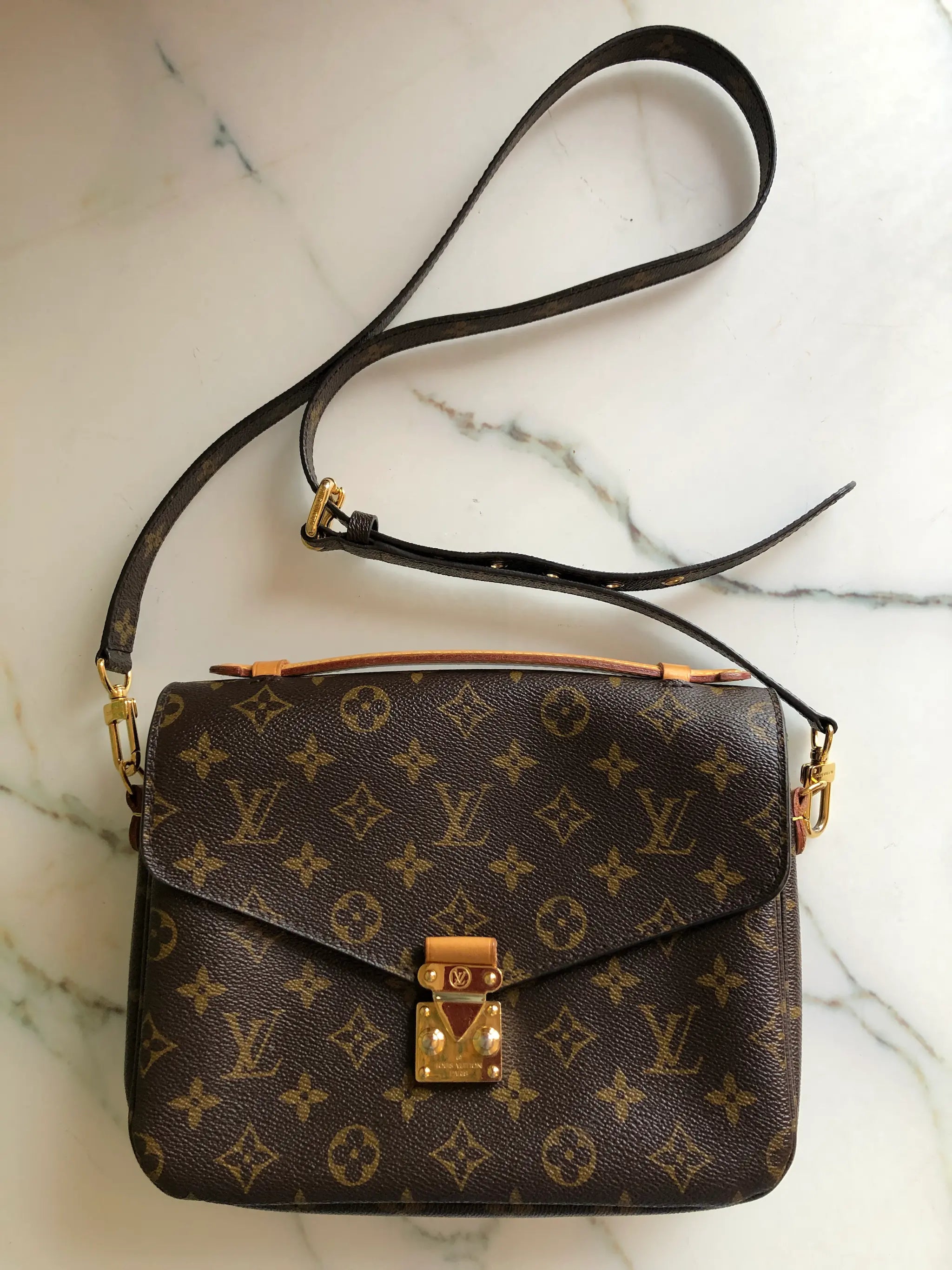 A Broochella Louis Vuitton Pochette Metis monogram shoulder bag in brown coated canvas with gold-tone hardware, resting on a white marble surface. The bag features the classic LV monogram pattern, a top flap with a gold-tone padlock closure, and an adjustable black leather shoulder strap. The background is white marble