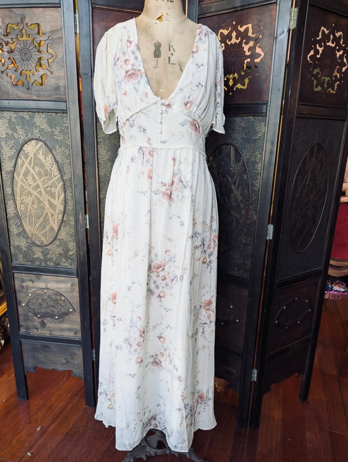 A Broochella LoveShackFancy White silk maxi dress in extra small, with pale pink and blue floral print. It is pictured on a mannequin in front of decorative wooden panels.