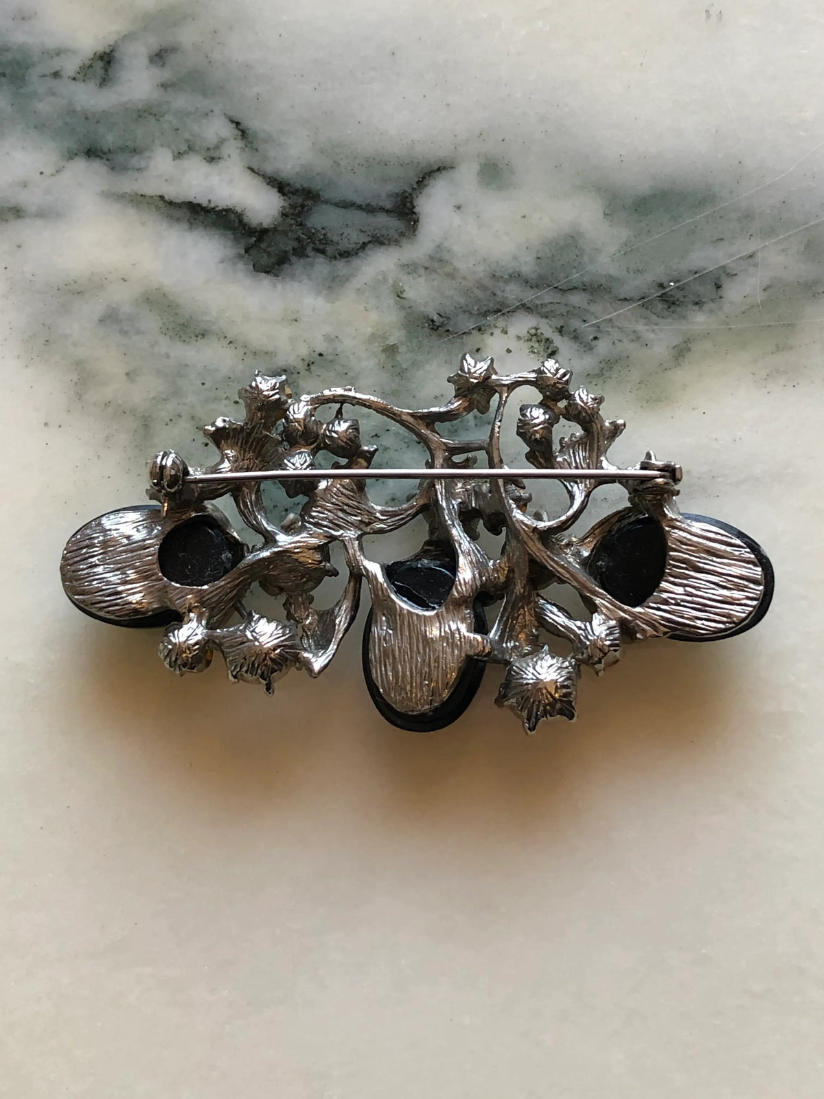 Broochella Luciana Vintage Black Cabochon Floral Brooch featuring three glossy black oval cabochons, clear crystals and sculptural silver-tone leaves on a cream and grey marble background.