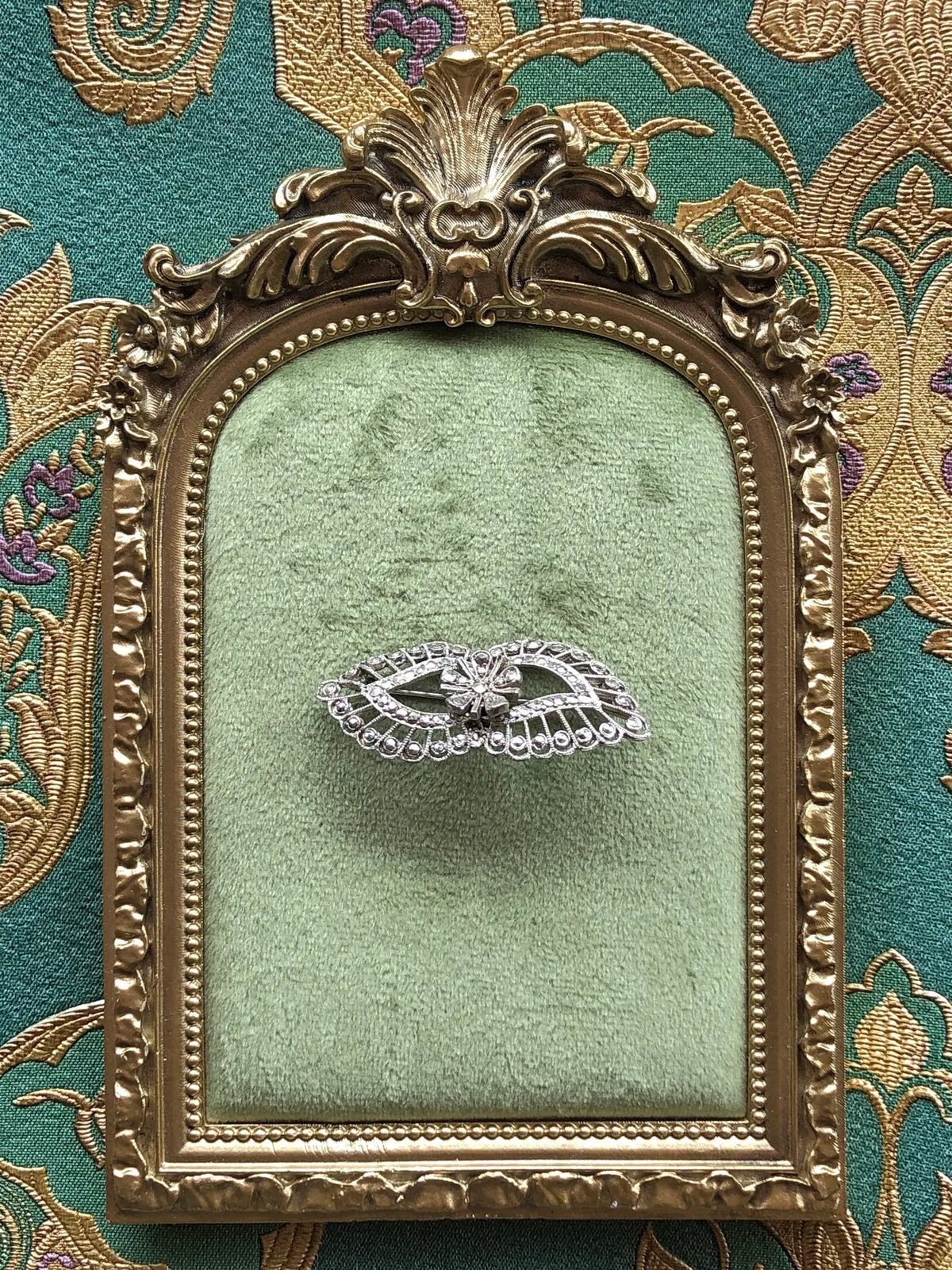 Broochella Luciana Vintage Silver Filigree Crystal Brooch featuring an elegant openwork design with sparkling clear crystals and a central floral motif, displayed on sage green velvet inside an ornate antique gold frame against green and gold embroidered fabric.