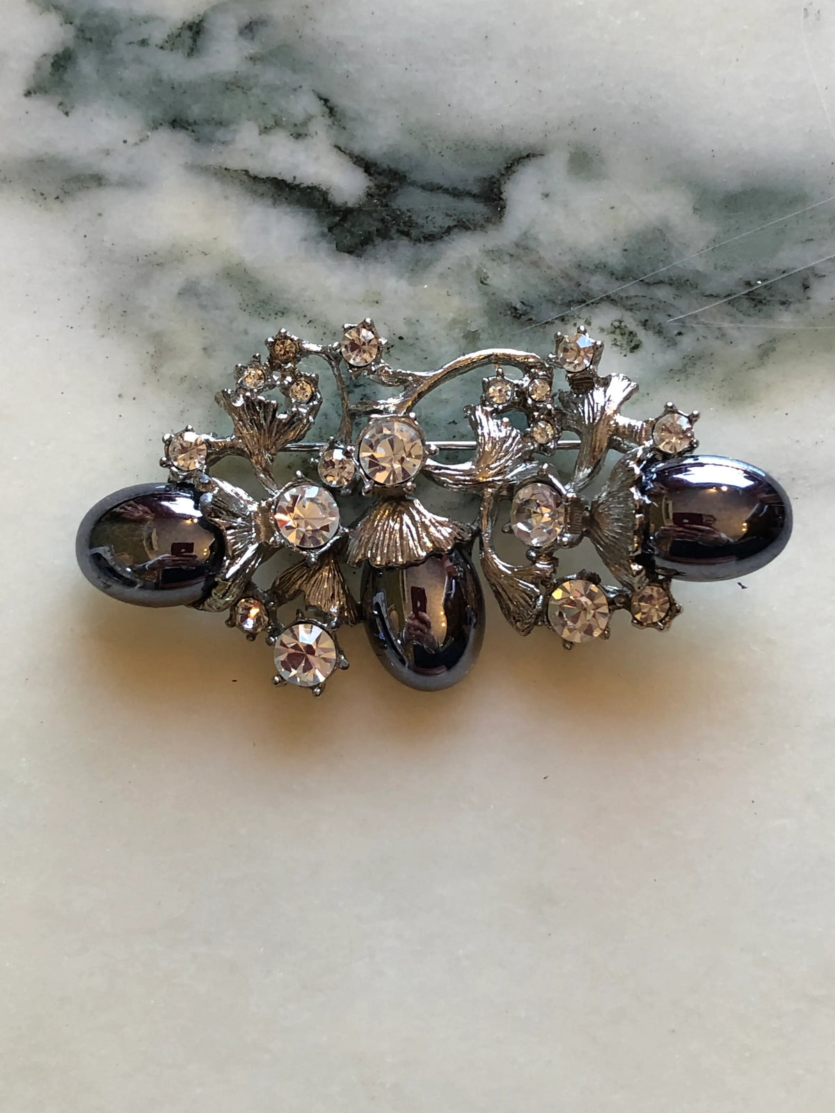 Broochella Lucilla Vintage Gunmetal Cabochon Floral Brooch featuring three polished gunmetal oval cabochons, clear crystals and sculptural silver-tone leaves on a cream and grey marble background.