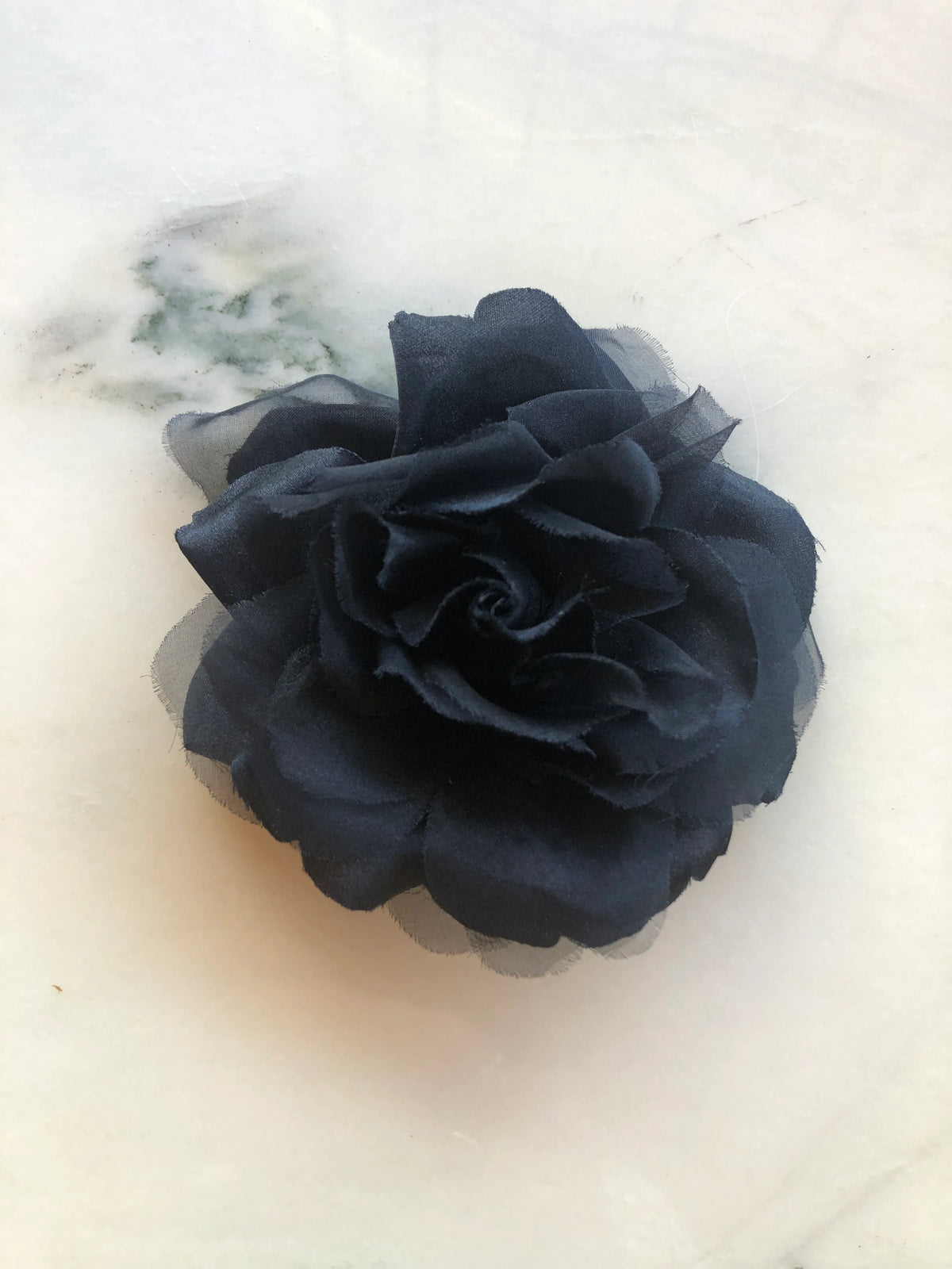 Broochella Ludovica black flower brooch with layered petals and sheer edges on a pale marble background.