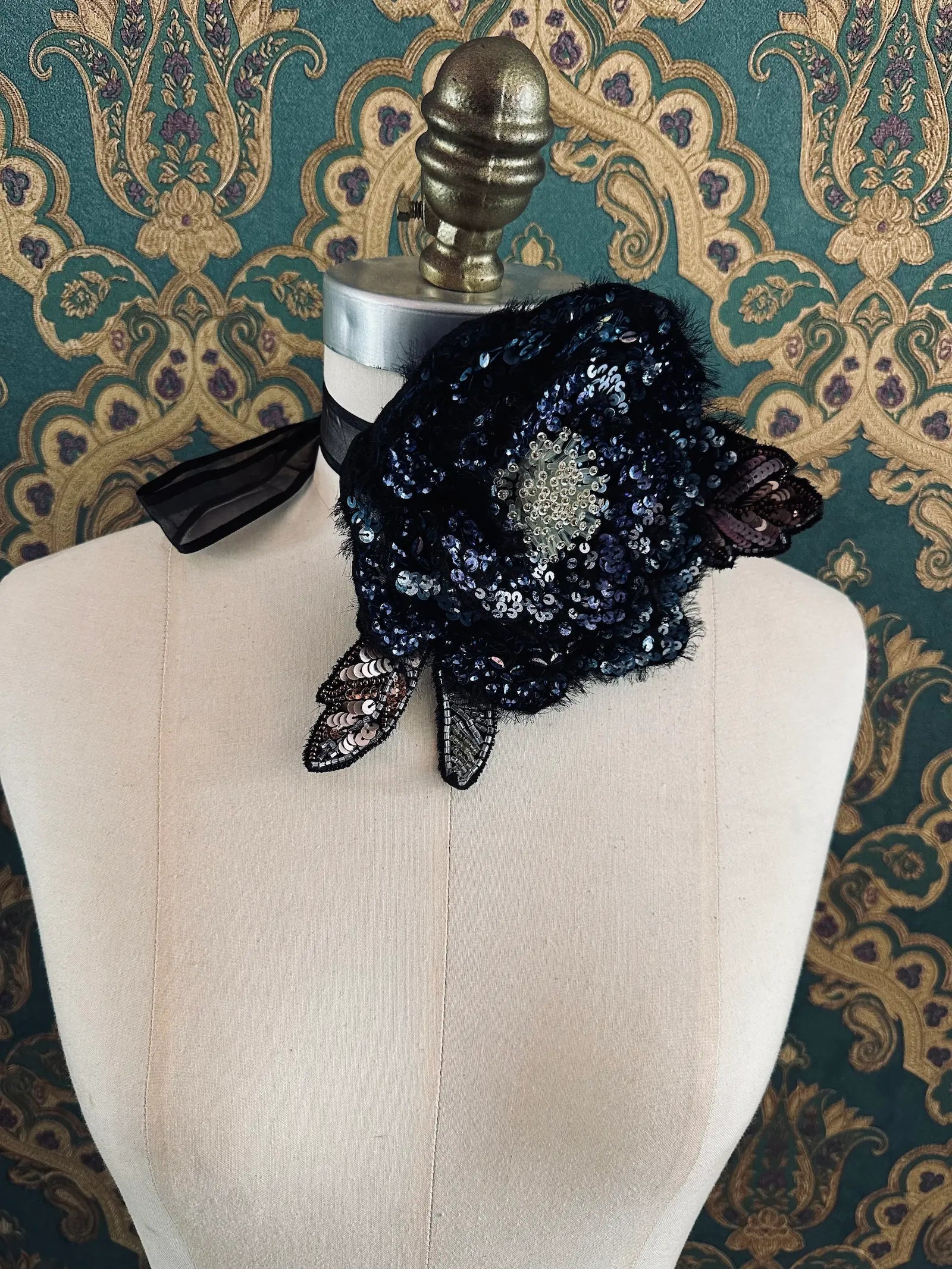 A Broochella luminosa  black and blue sequinned floral choker and brooch with lace leaves and a sheer black ribbon bow, displayed on a cream-coloured dress form against a green and gold patterned wallpaper background.