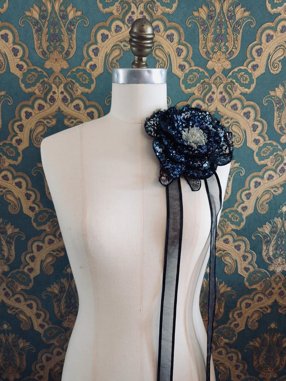 A Broochella luminosa  black and blue sequinned floral choker and brooch with lace leaves and a sheer black ribbon bow, displayed on a cream-coloured dress form against a green and gold patterned wallpaper background.