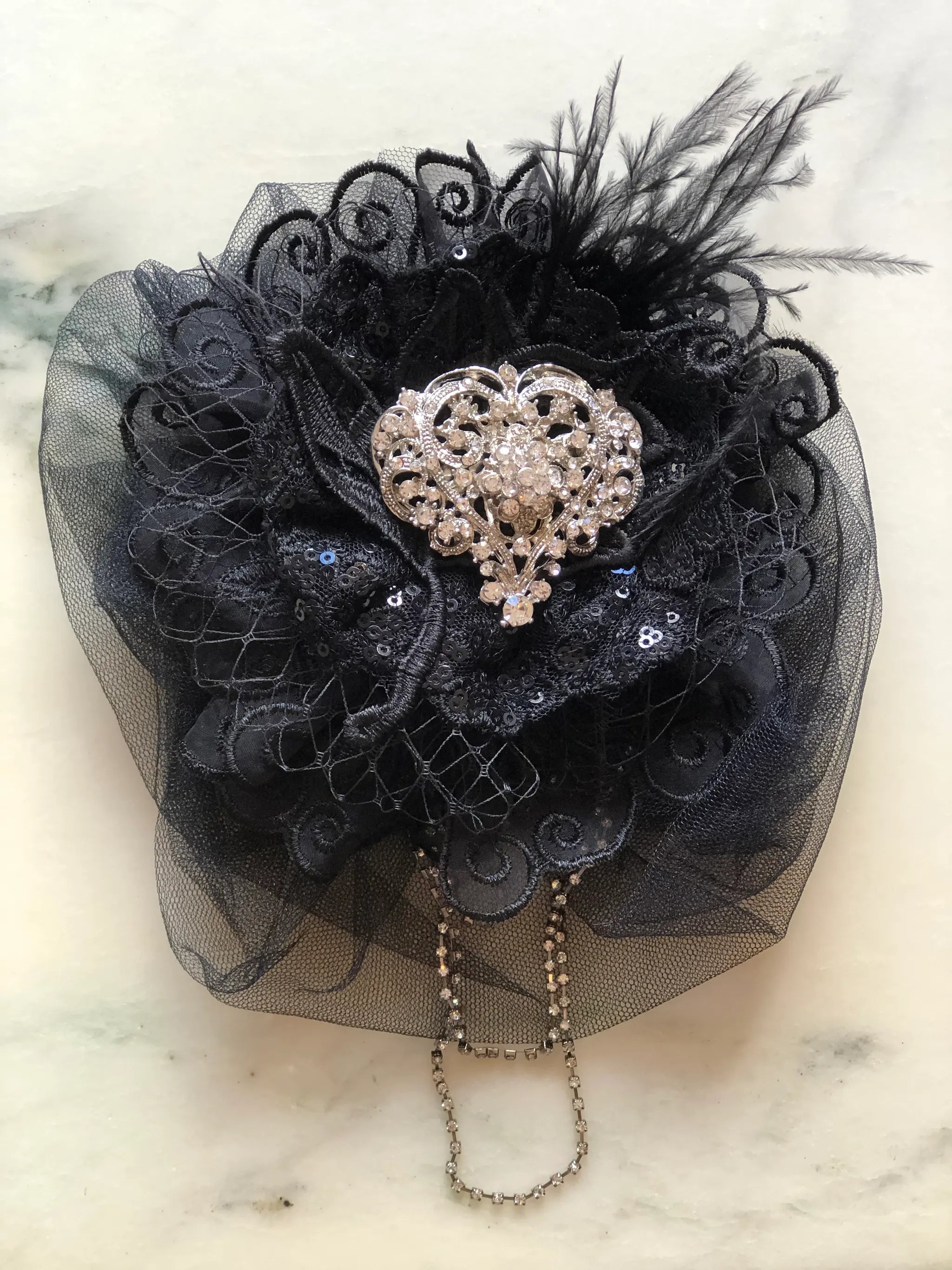 A Broochella Madonna black handmade lace brooch, with a central silver rhinestone brooch, black feathers, and a dangling silver chain, displayed on a light marble surface.