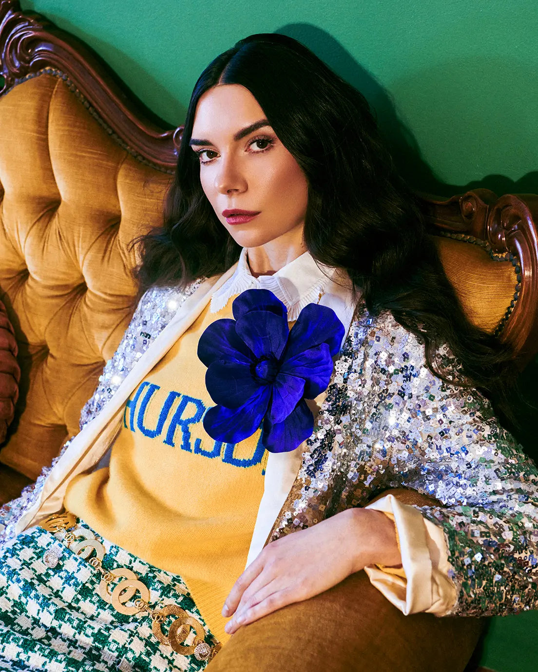 Woman sitting on a vintage couch wearing a yellow sweatshirt with a Broochella purple magnolia flower brooch and sequin jacket.