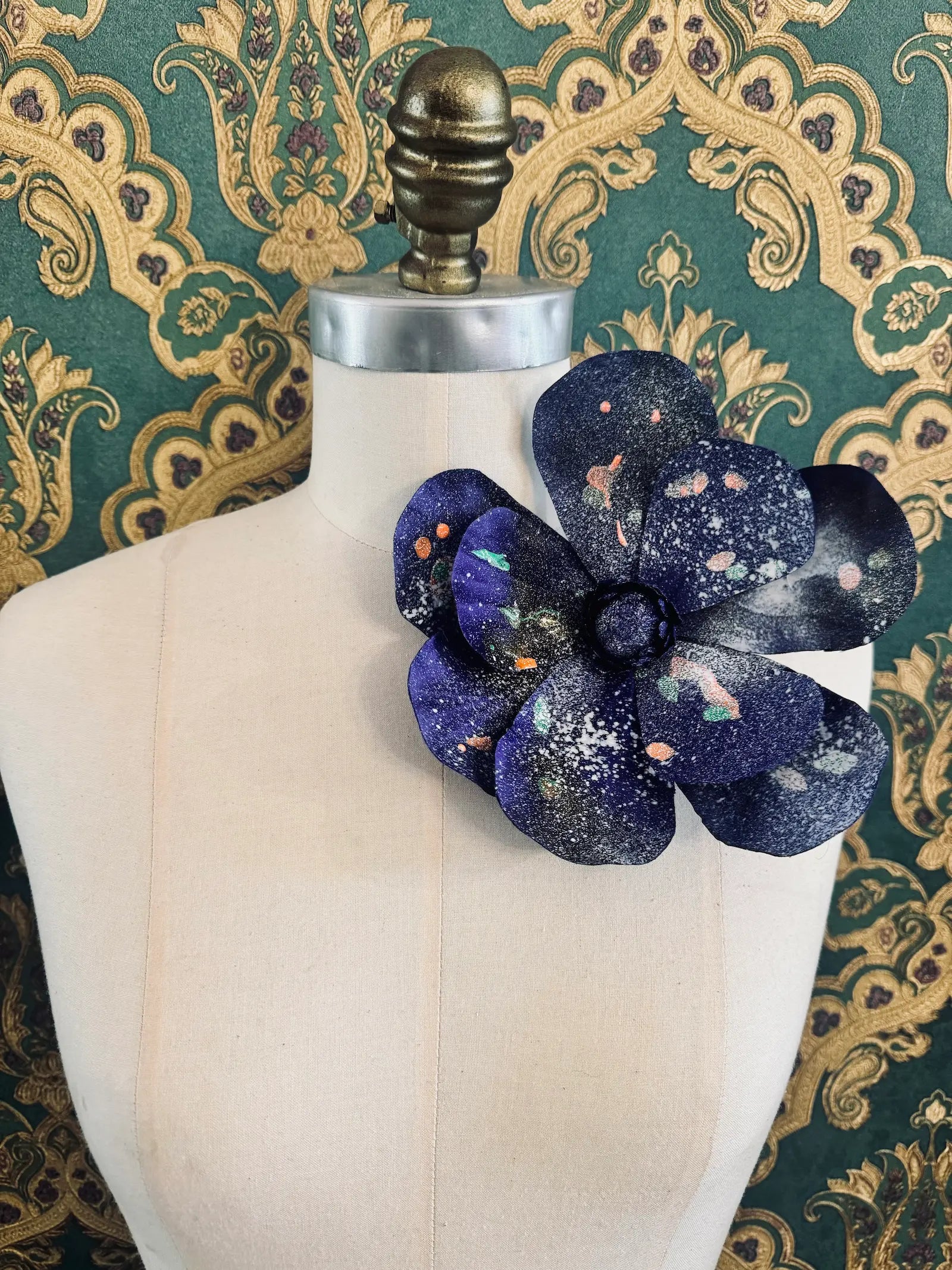 Broochella IMagnolia d'arte large blue flower brooch with multi coloured paint splatter. One of a kind customized piece. Pictured on a mannequin.  The background is green and gold damask wallpaper.