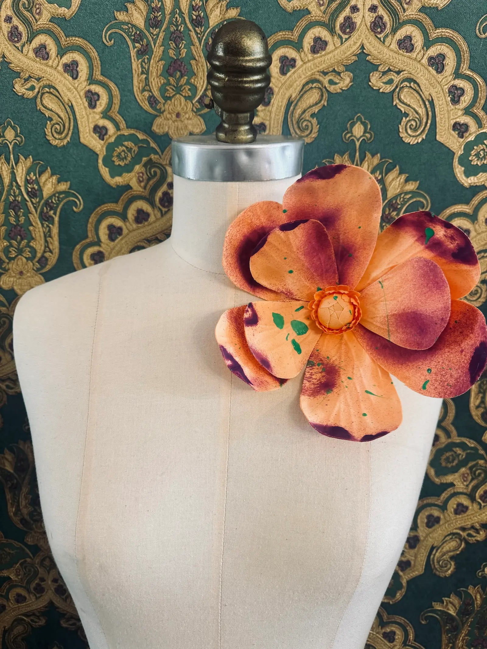 Broochella IMagnolia d'arte large orange flower brooch with multi coloured paint splatter. One of a kind customized piece. Pictured on a mannequin.  The background is green and gold damask wallpaper.