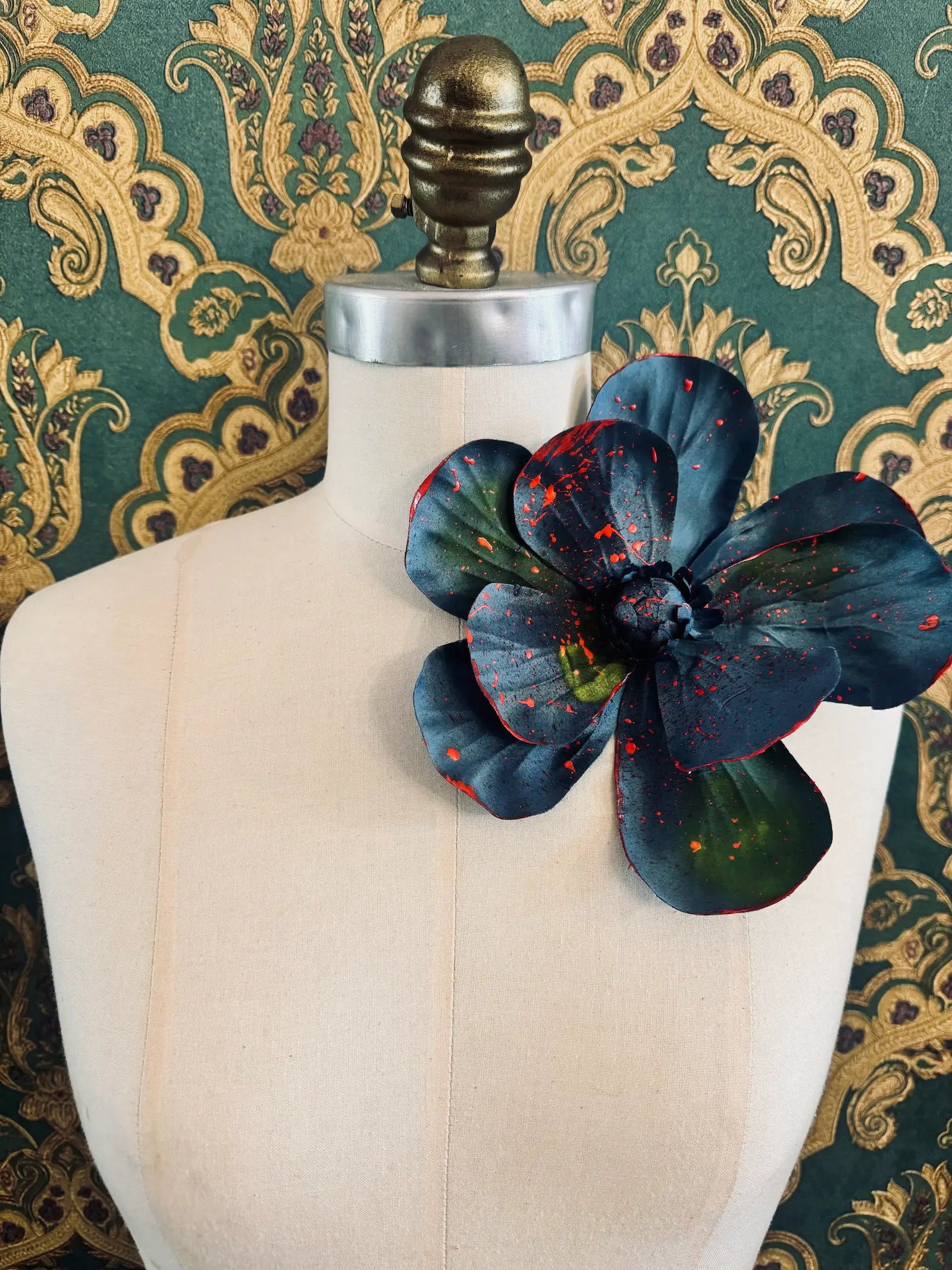 Broochella IMagnolia d'arte large blue flower brooch with multicolour paint splatter. One of a kind customized piece. Pictured on a mannequin.  The background is green and gold damask wallpaper.