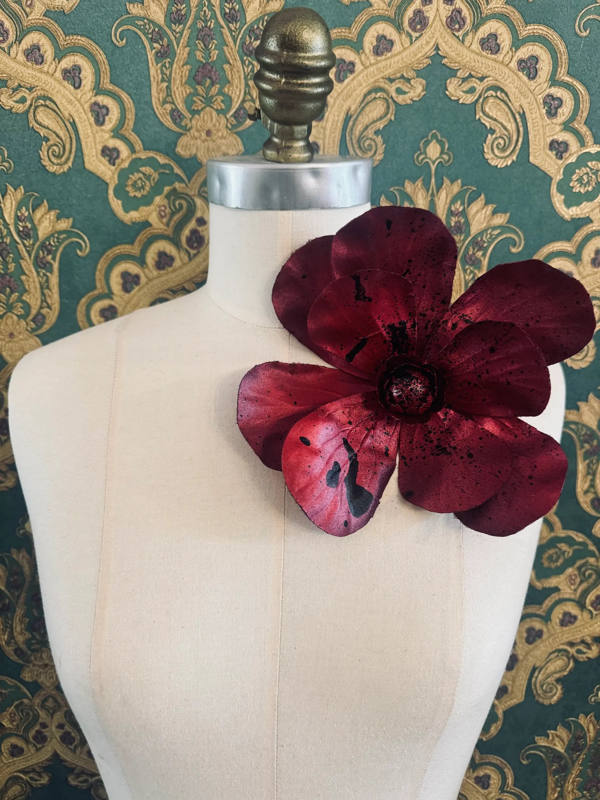 Broochella IMagnolia d'arte large red flower brooch with black paint splatter. One of a kind customized piece. Pictured on a mannequin.  The background is green and gold damask wallpaper.