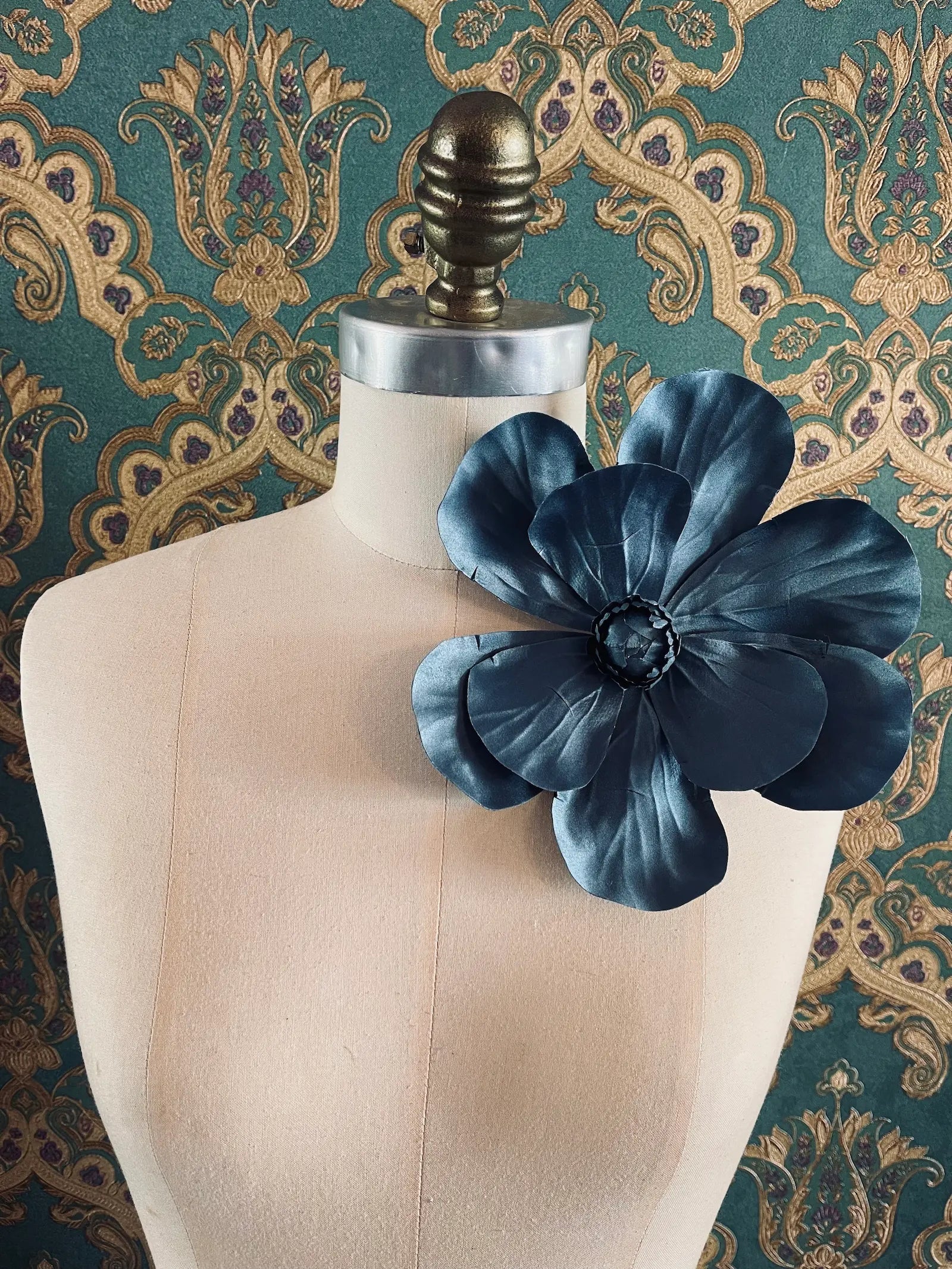 Broochella Magnolia handmade large flower brooch in blue. Styled on a mannequin. The background is green and gold damask wallpaper