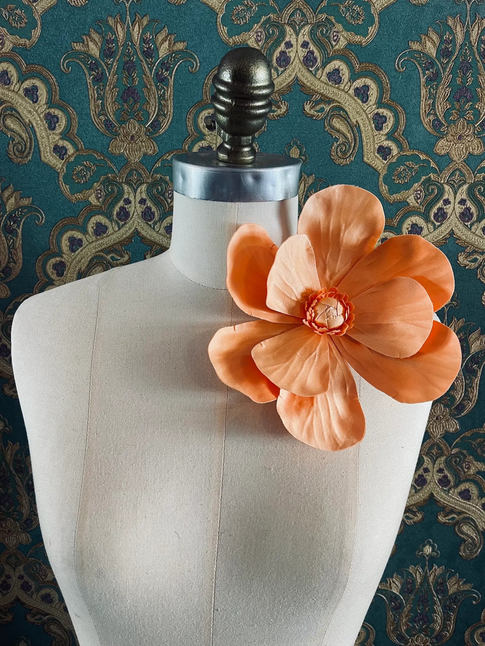 Broochella Magnolia handmade large flower brooch in orange. Styled on a mannequin. The background is green and gold damask wallpaper