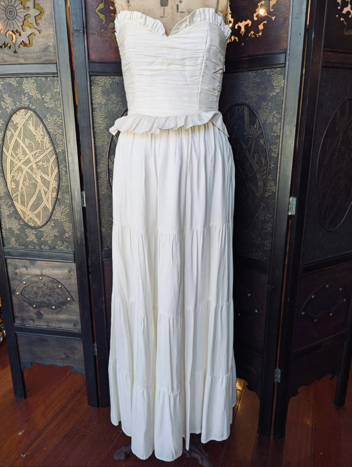 Broochella Malie Ivory Strapless Maxi Dress with gathered bodice, small ruffle peplum and tiered maxi skirt. It is pictured on a mannequin. The background is an ornate, dark wood room divider. 