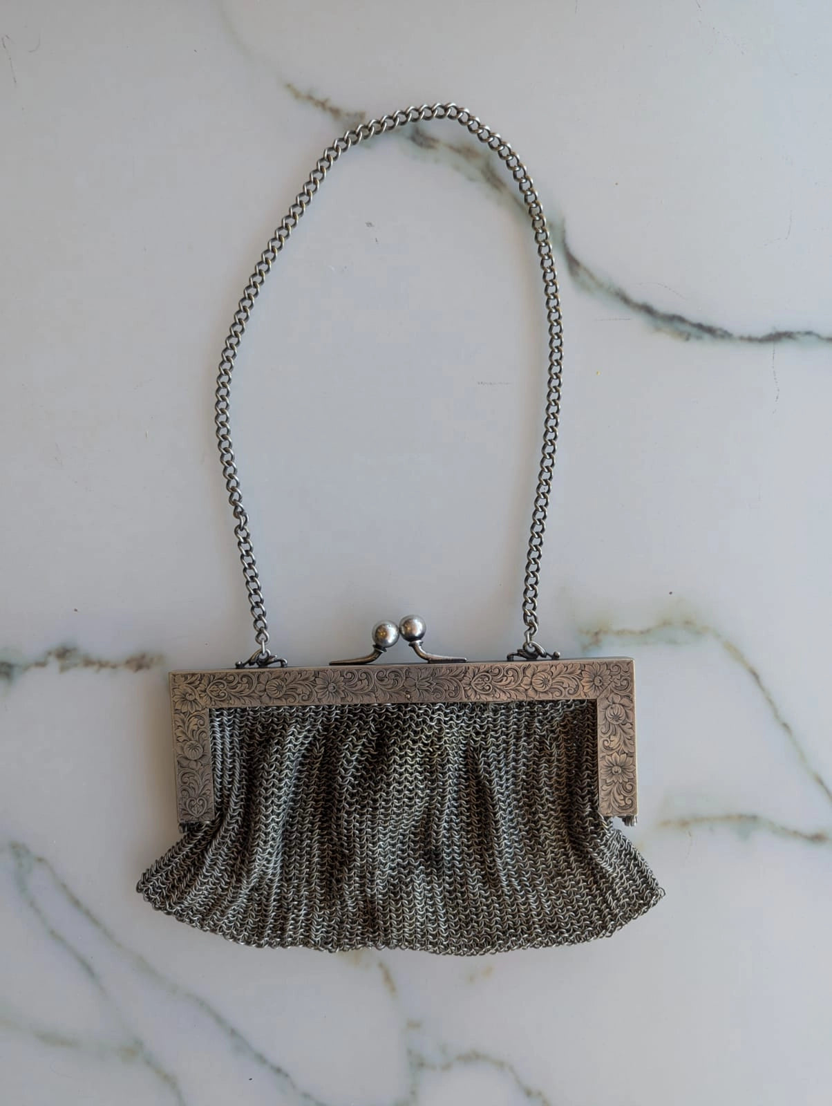 A vintage Broochella maison broche bijou argente silver chain mesh clutch purse with a textured silver-tone frame, brooch embellishment , and dangling pearl tassel, displayed on a white marble background.