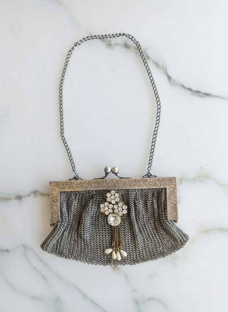 A vintage Broochella maison broche bijou argente silver chain mesh clutch purse with a textured silver-tone frame, brooch embellishment , and dangling pearl tassel, displayed on a white marble background.