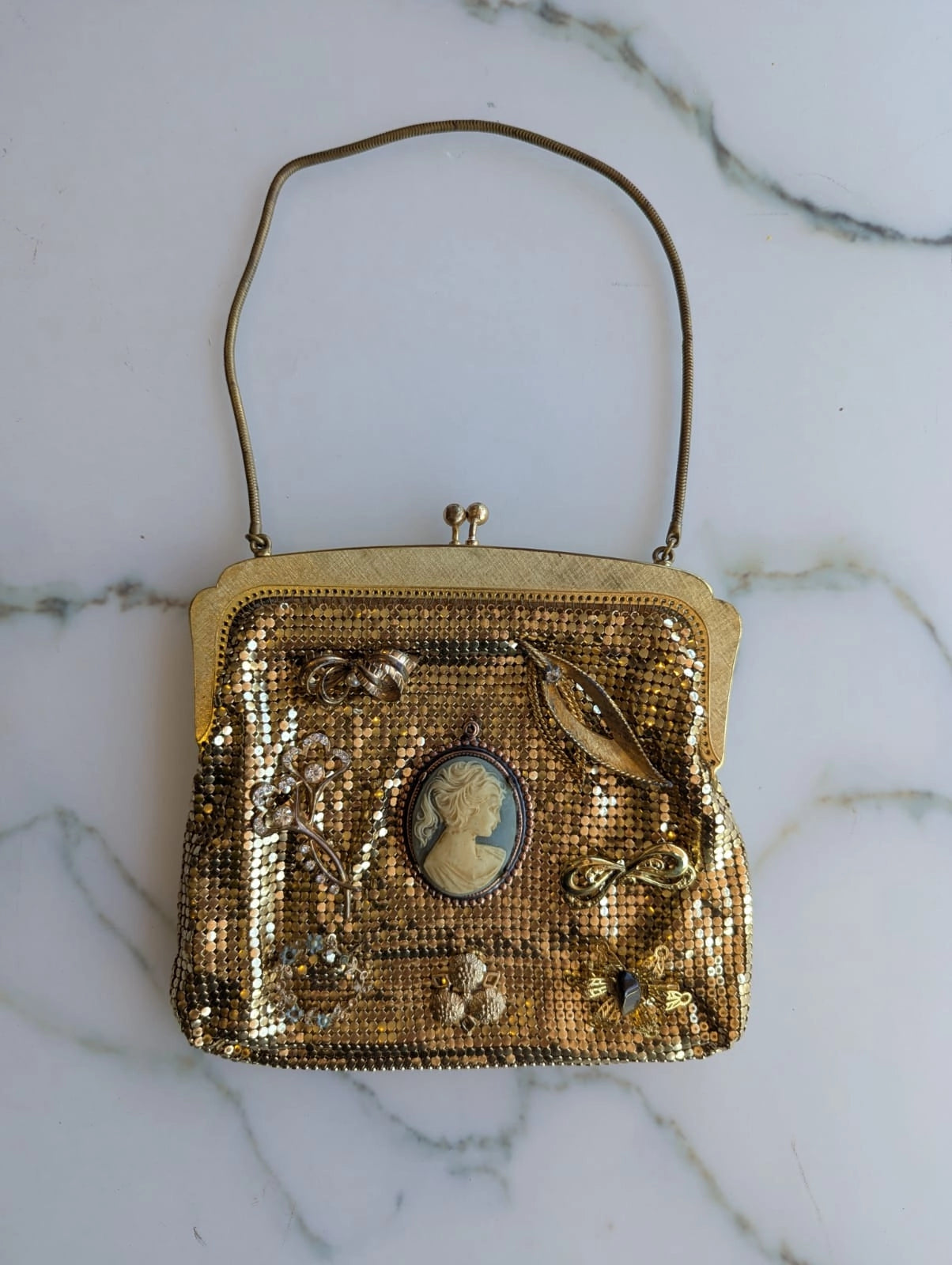 A Broochella etoile d'or embellished vintage gold-tone mesh Oroton clutch purse with a curved shoulder strap, featuring a central cameo-style oval portrait, floral embellishments, and a bird motif, displayed on a white marble surface.