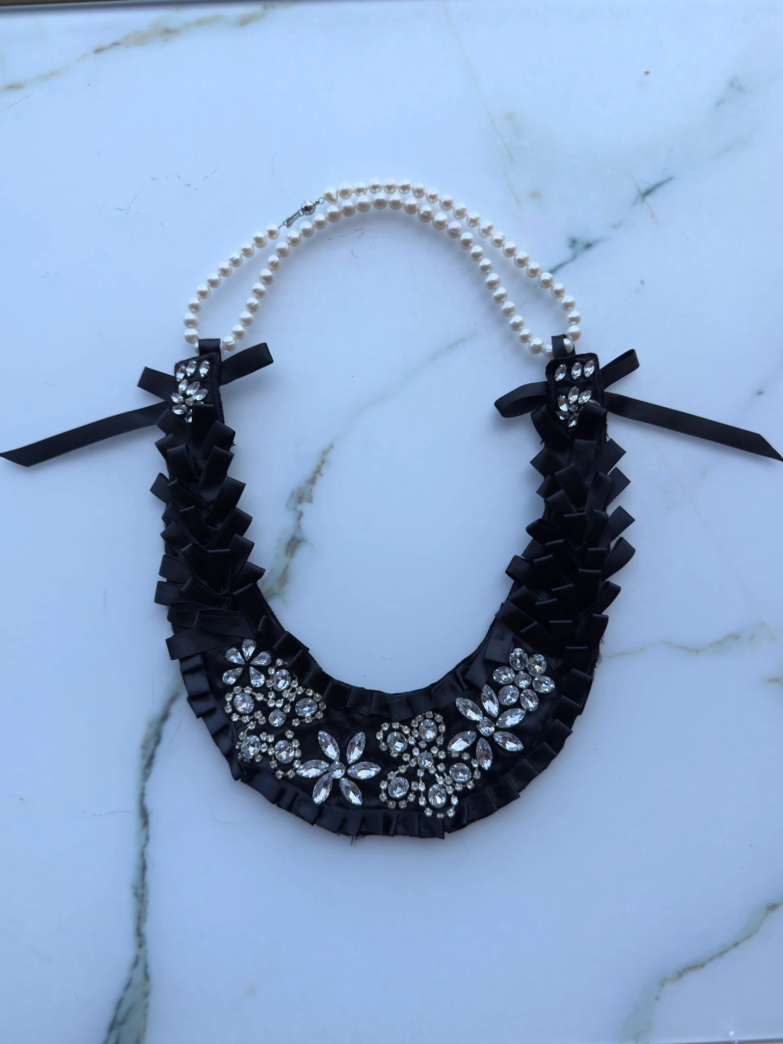 Broochella Maison Broche l'amour pare handmade black satin decorative beaded necklace/bib with clear crystal embellishment. A pearl necklace is added to create a neckpiece. Pictured on a white marble table