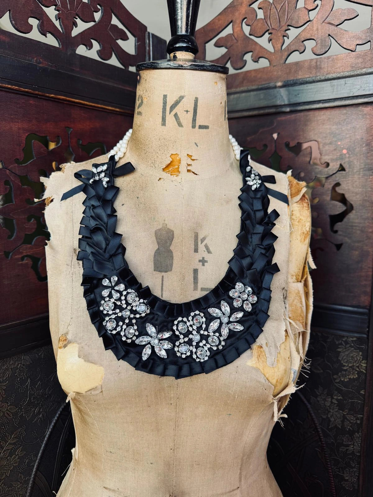 Broochella Maison Broche l'amour pare handmade black satin decorative beaded necklace/bib with clear crystal embellishment. It is presented on a vintage mannequin. The background is a brown, wooden decorative screen. 